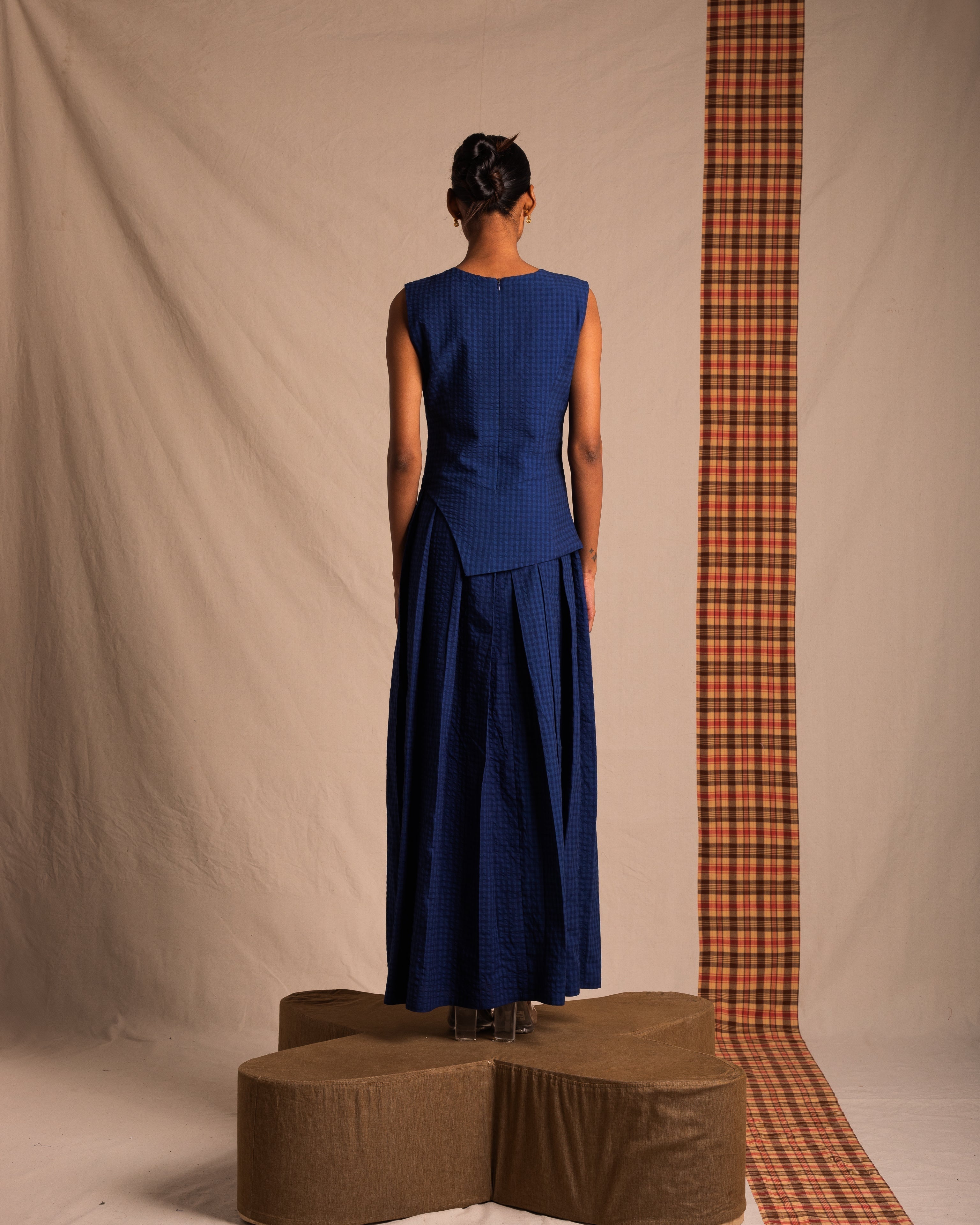 Woman in sleeveless blue maxi dress standing on brown platform with beige and plaid backdrop