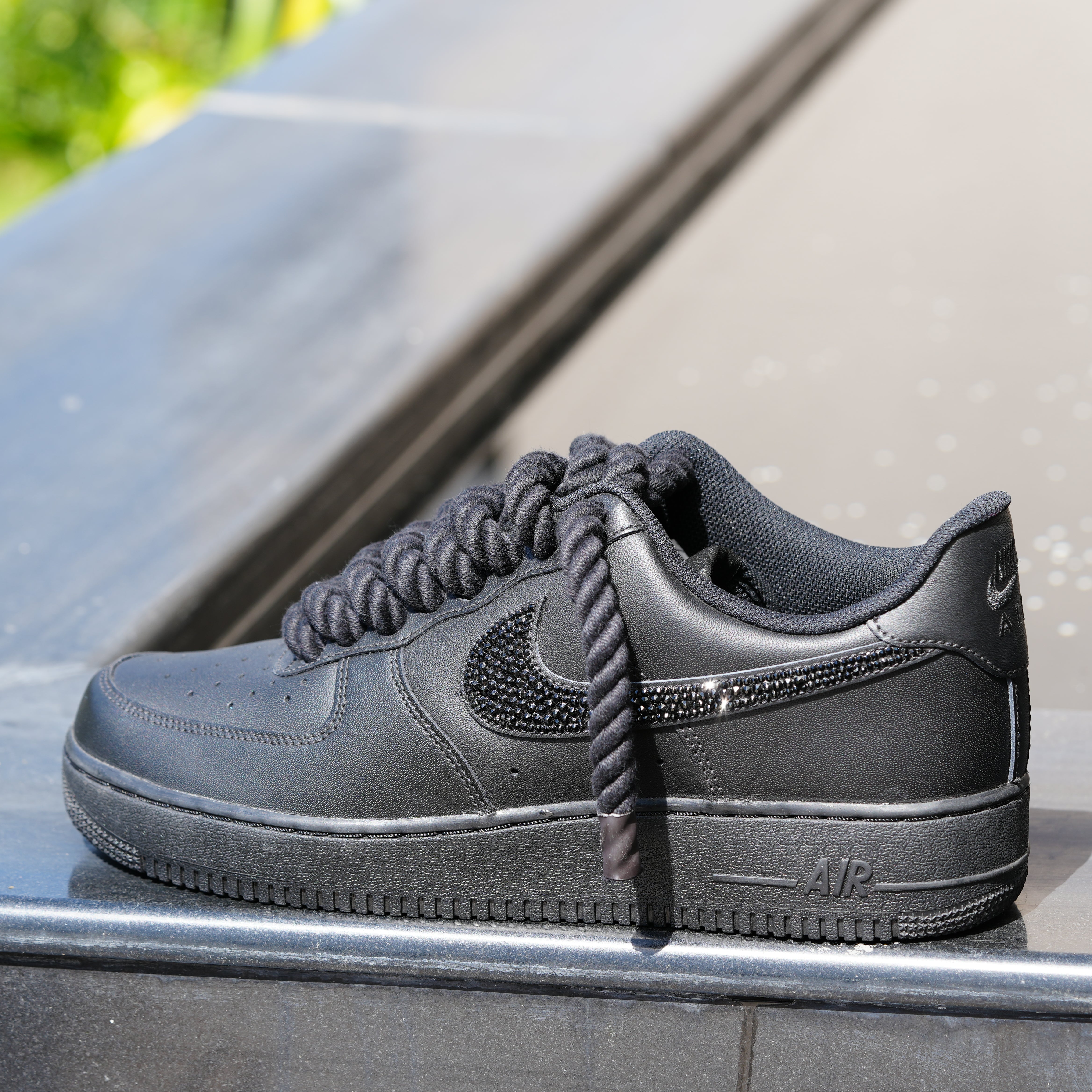 Black Nike Air Force 1 sneaker with textured laces and jeweled Swoosh on outdoor ledge