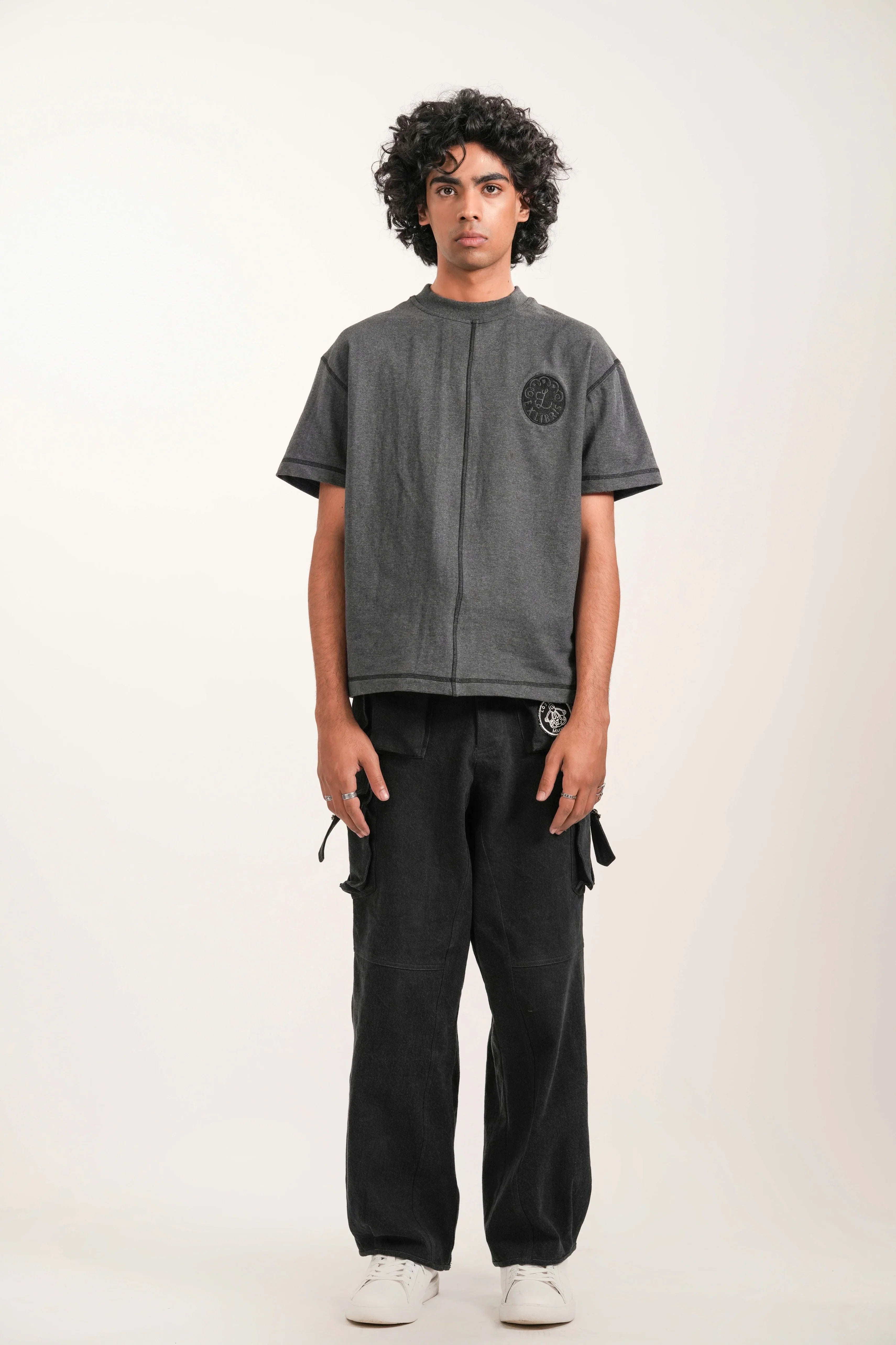 Young man standing in studio wearing gray embroidered t-shirt, black cargo pants, and white sneakers