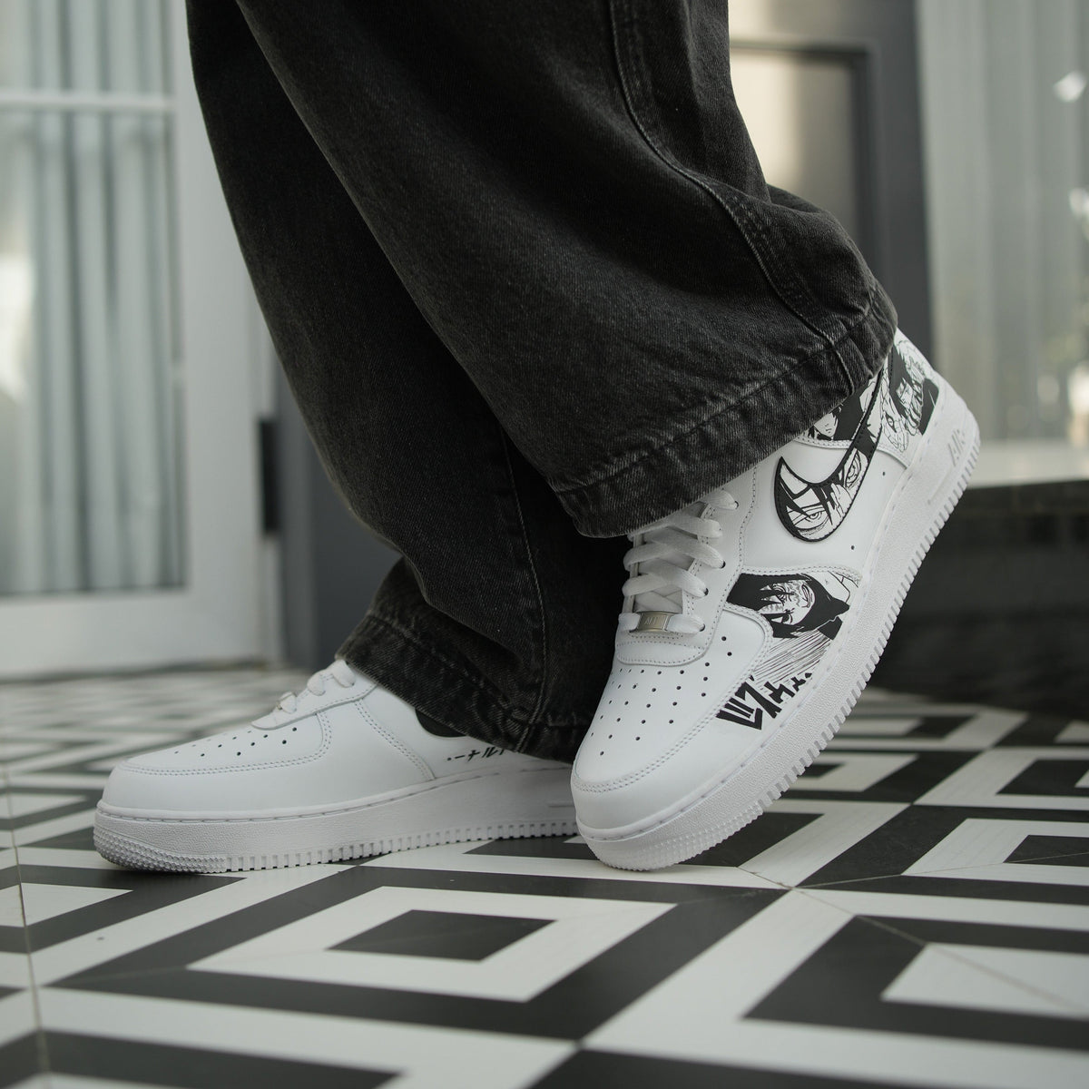 White streetwear sneakers with black graphic designs, worn with black jeans on patterned floor