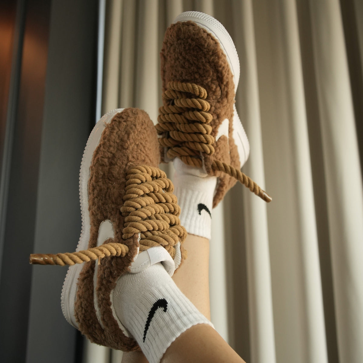 Brown fuzzy sneakers with thick rope laces, worn with white Nike socks, streetwear style.