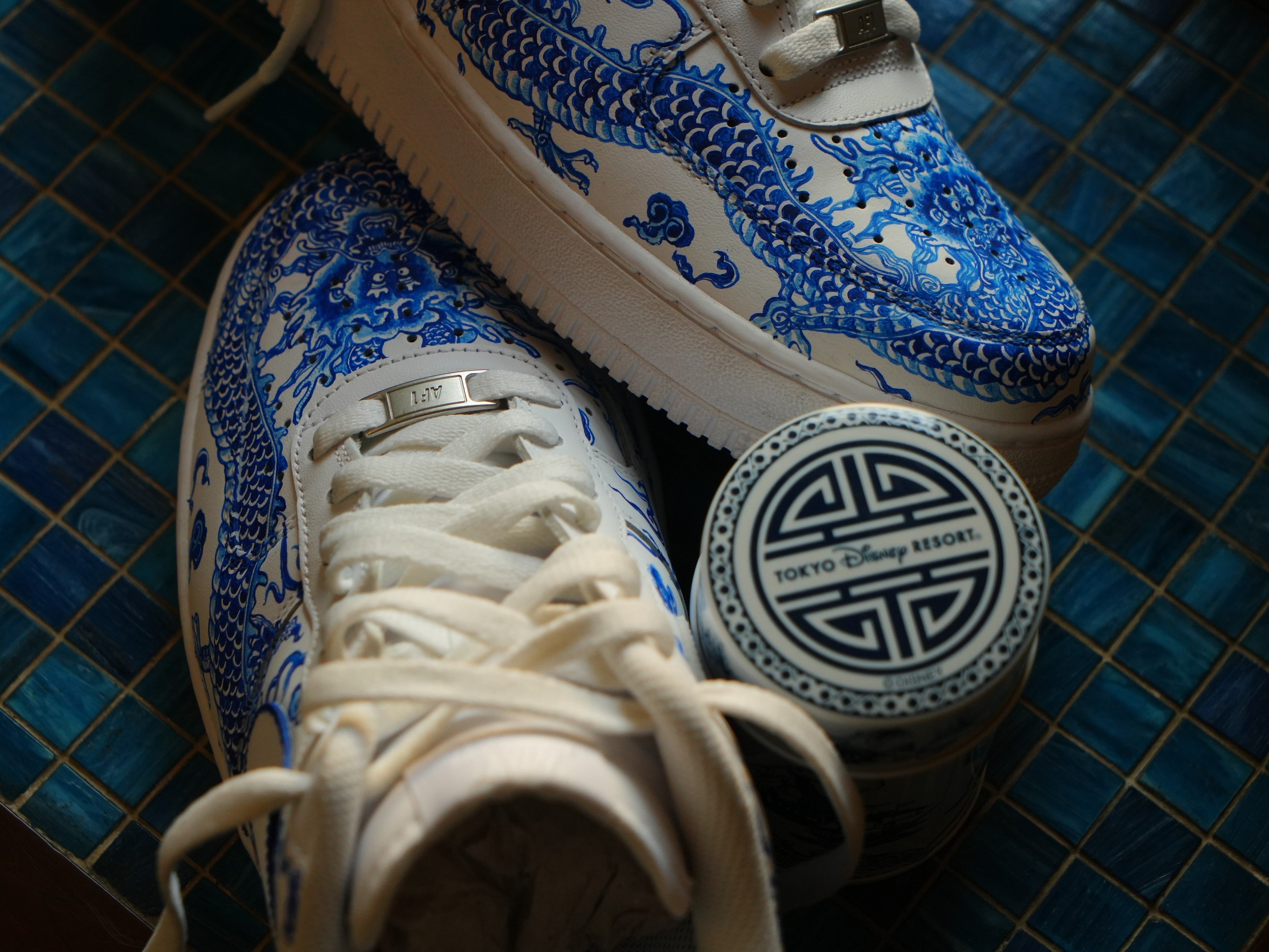 White sneakers with blue dragon design next to Tokyo Disney Resort tin on blue tile floor.