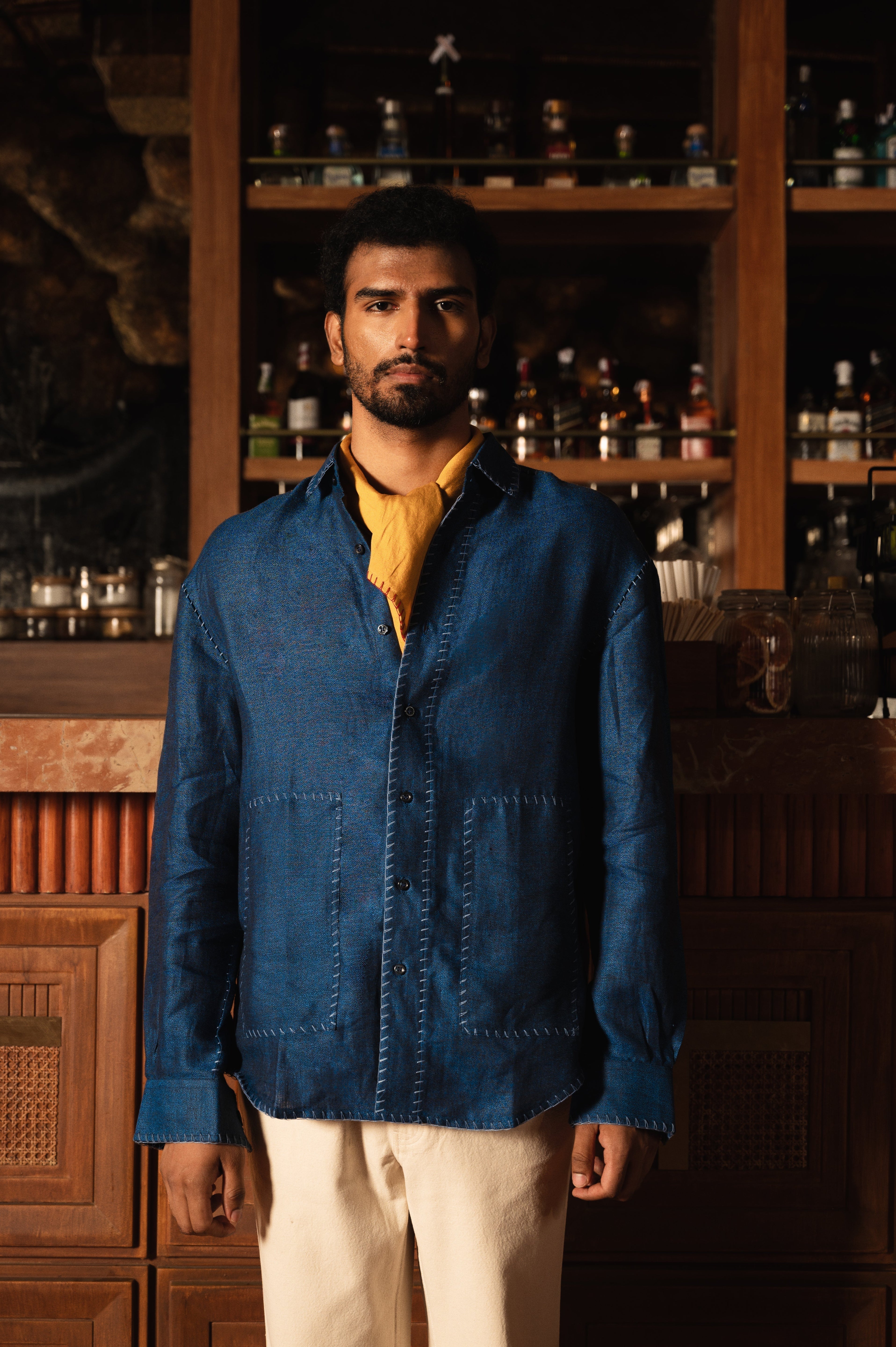 Man modeling blue streetwear button-up shirt with yellow scarf, standing indoors by bar shelves.