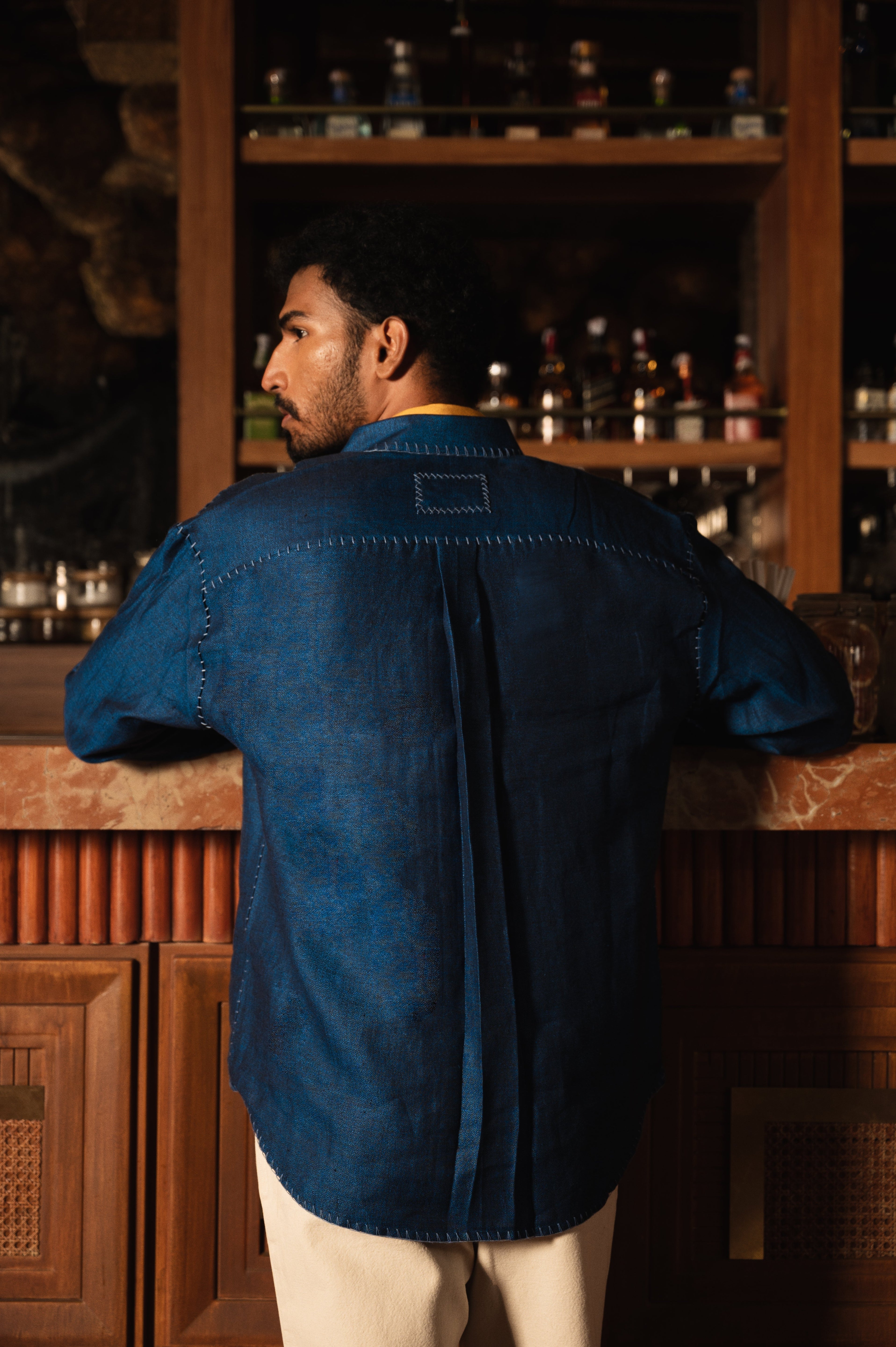 Man in a stylish blue stitched shirt and cream pants leaning at a bar counter