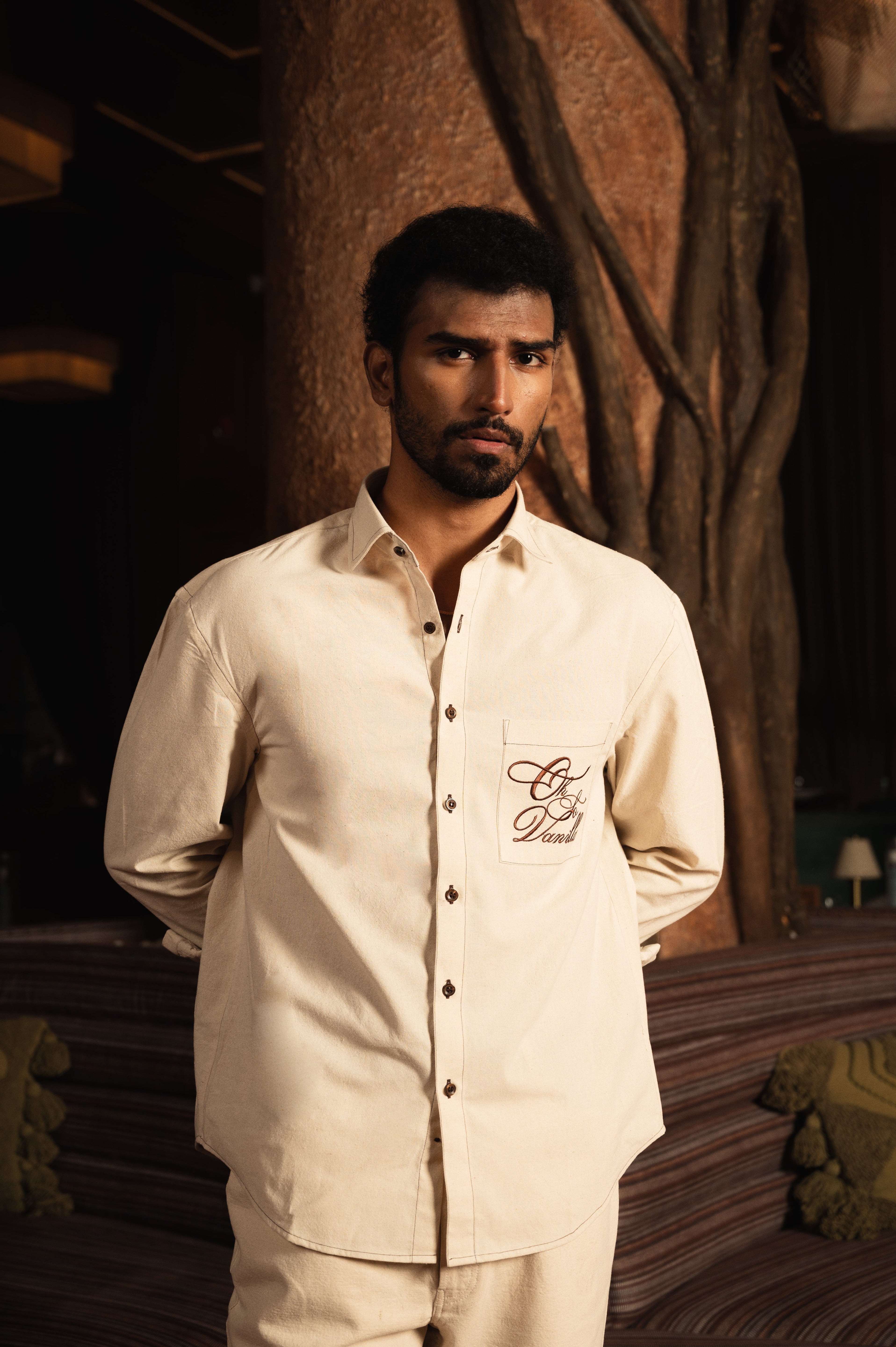 Man wearing a beige long sleeve streetwear shirt with embroidered script on pocket indoors