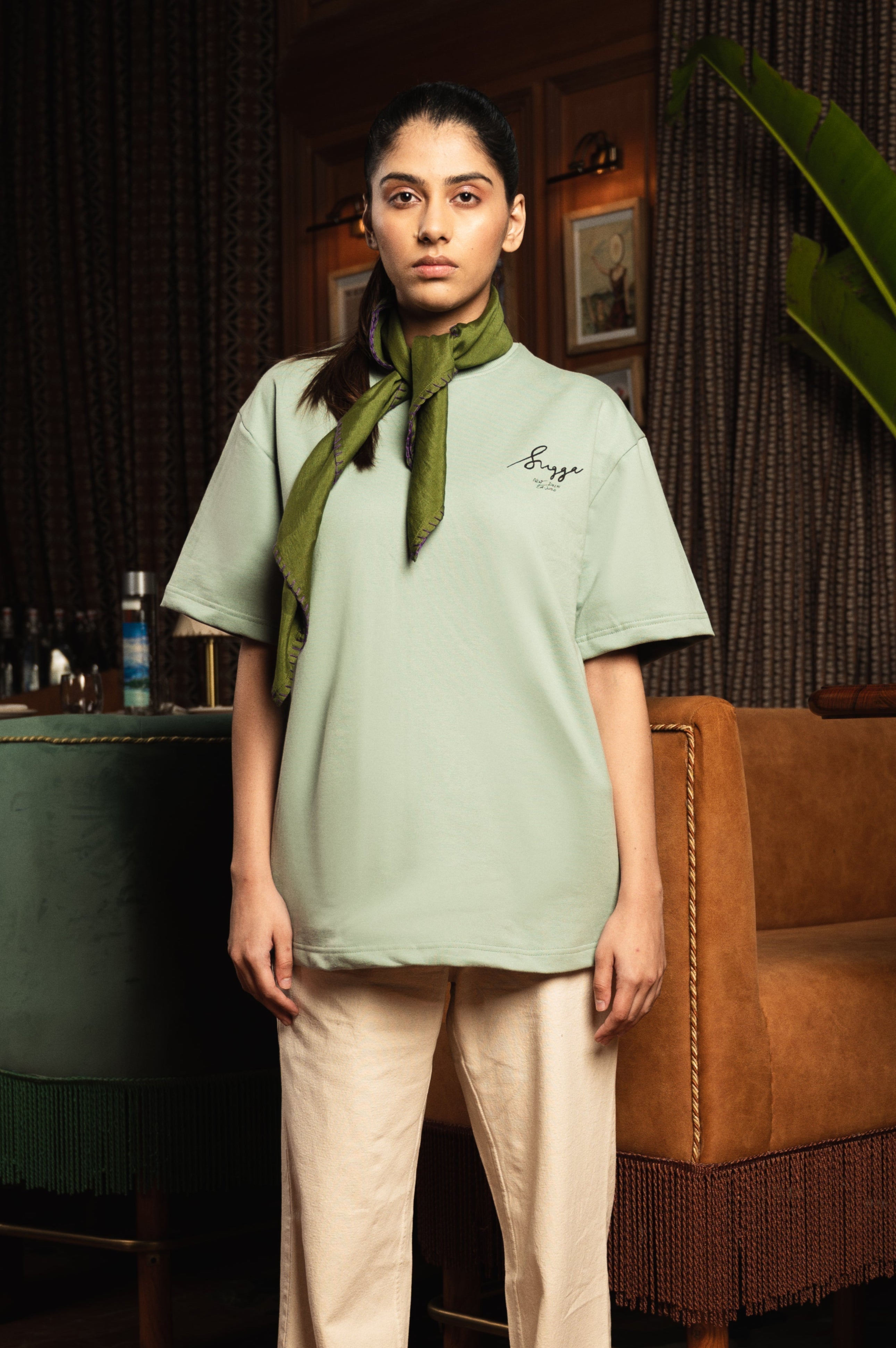 Woman in sage green oversized t-shirt with embroidered text, beige pants, and green scarf indoors