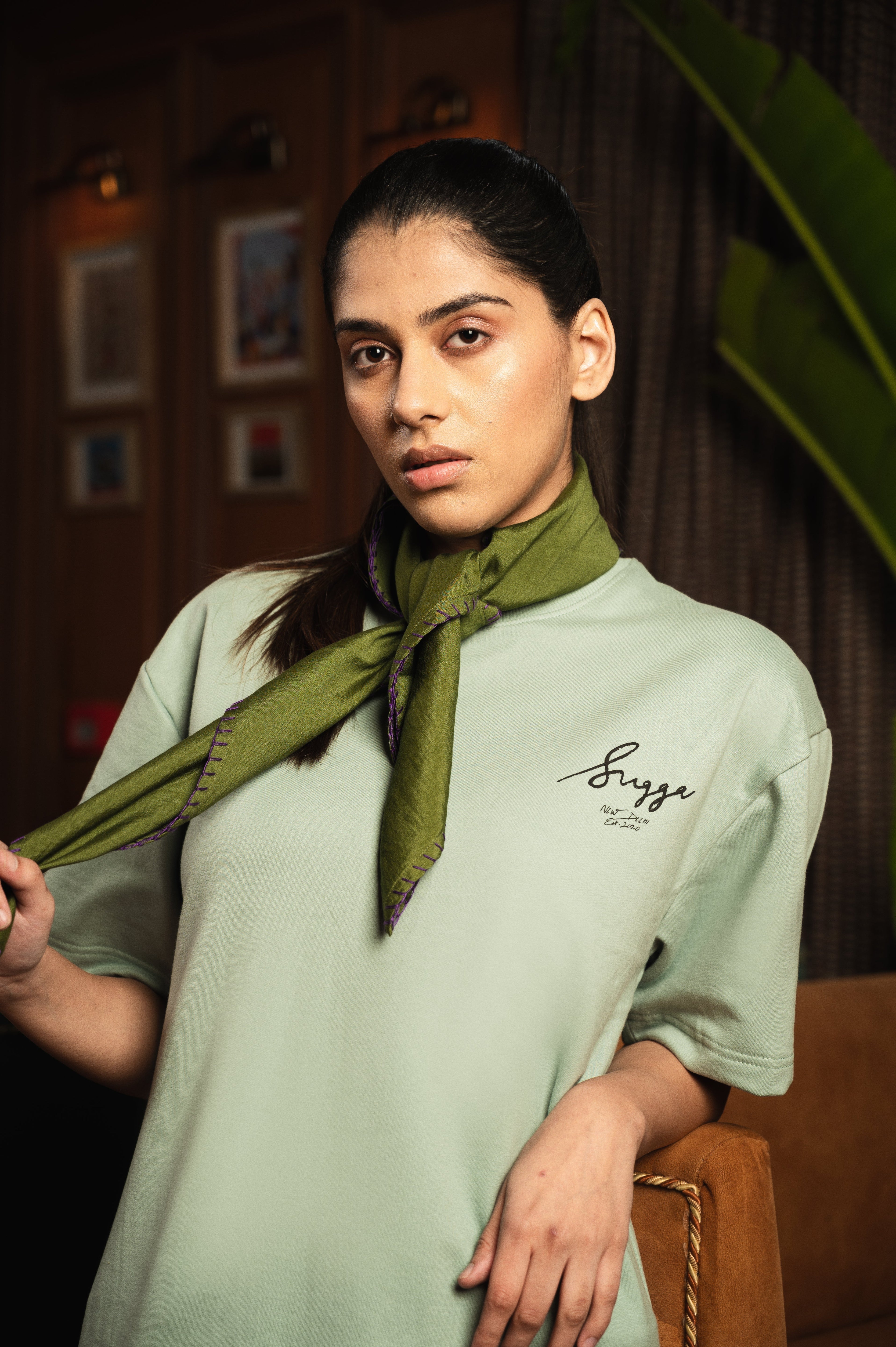 Woman in mint green streetwear tee with script logo, green scarf, indoors, plant background