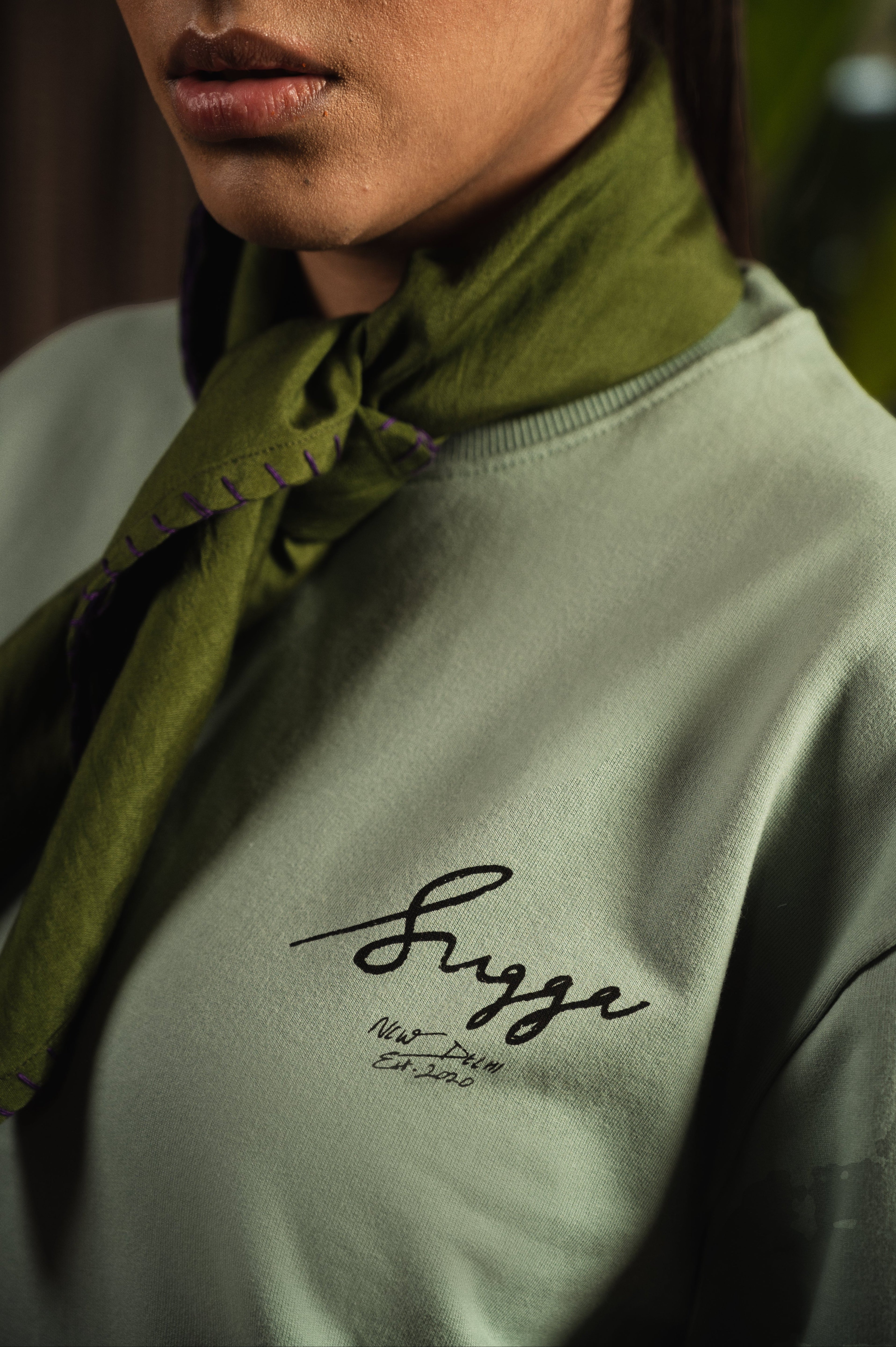 Close-up of a woman wearing a green streetwear sweatshirt with script logo and a tied scarf.