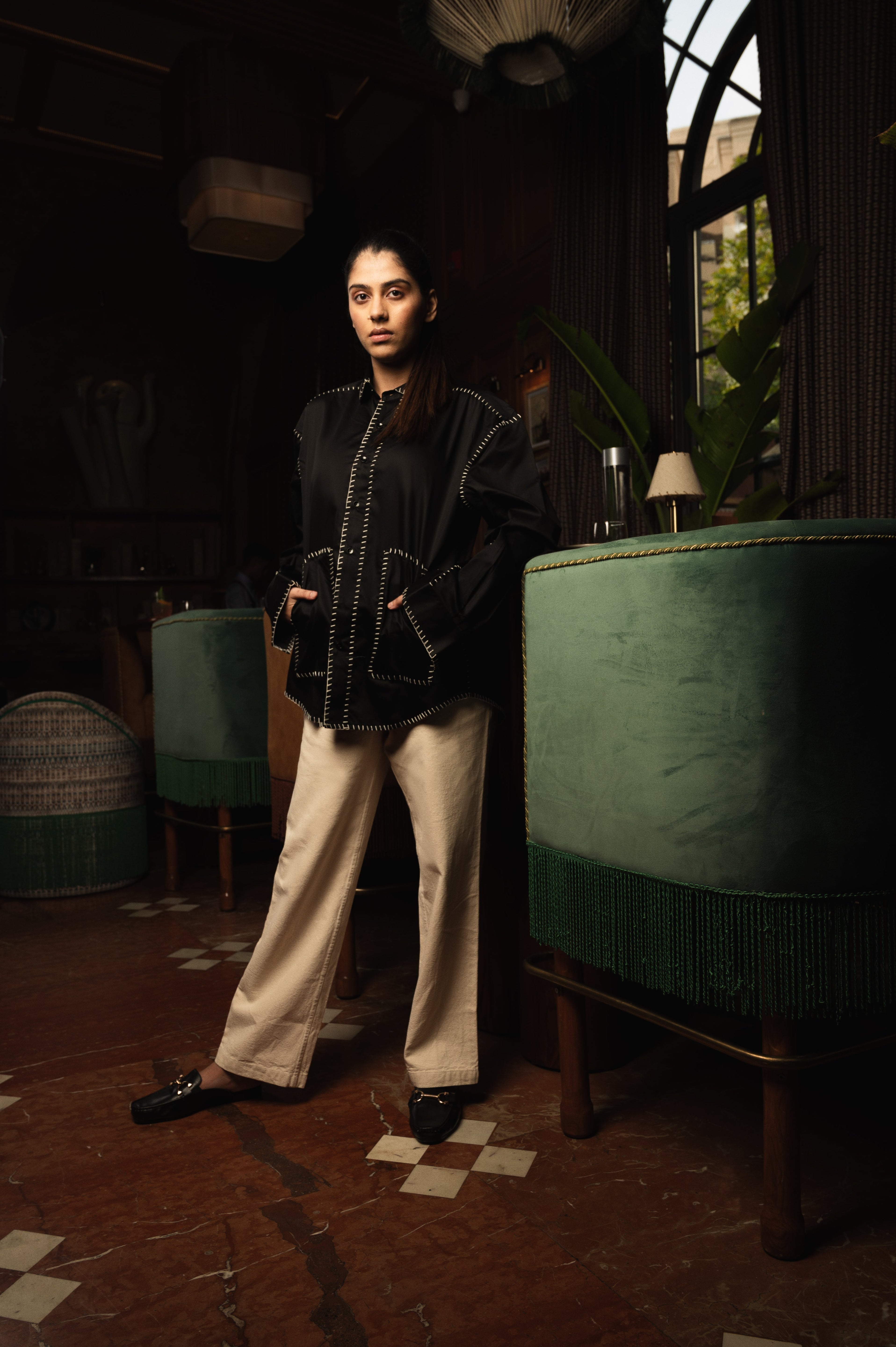 Woman in a black stitched jacket and beige pants, standing in a stylish lounge with green decor.