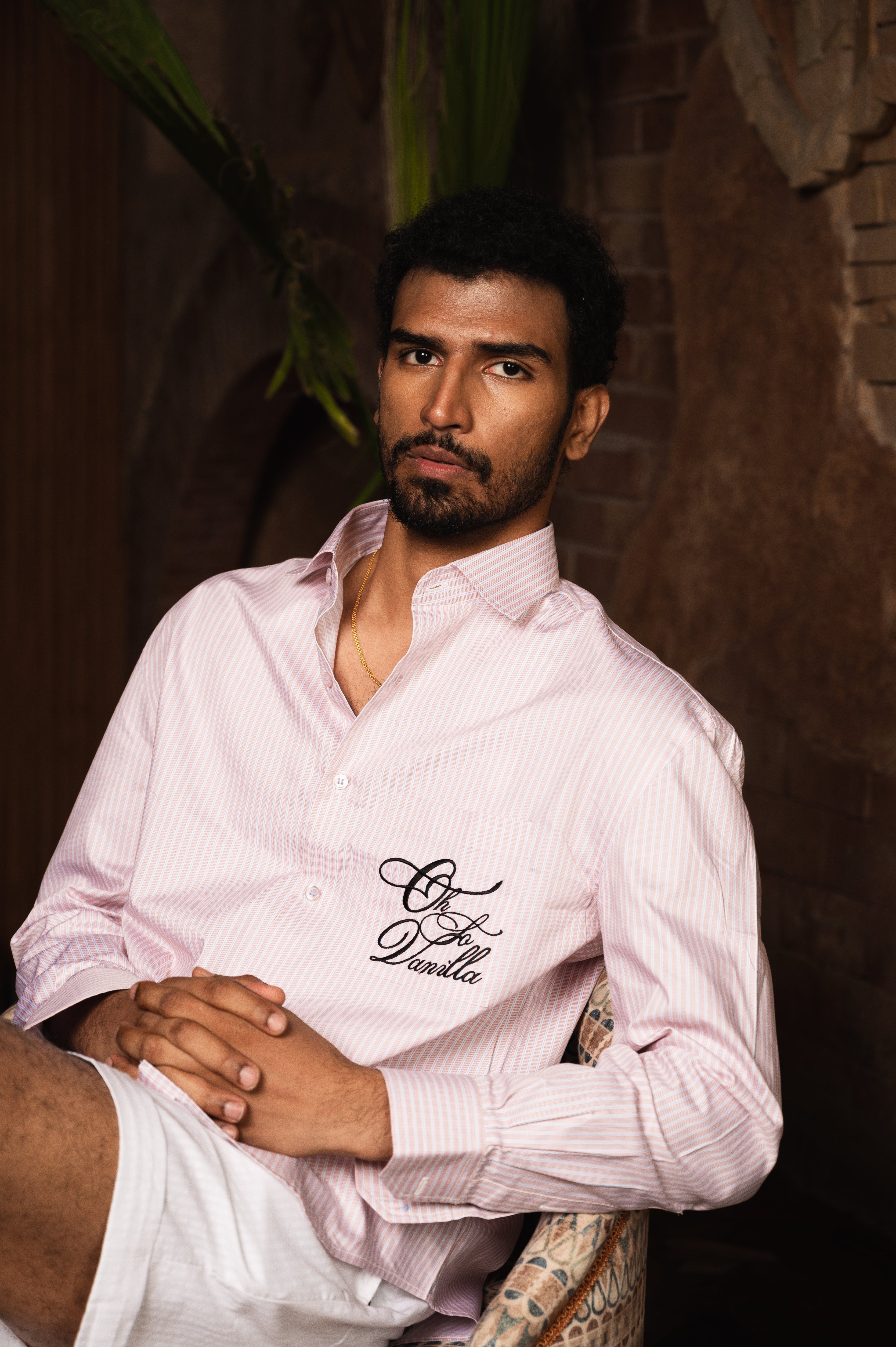 Man in pink striped streetwear shirt with embroidered text, seated indoors, luxury fashion