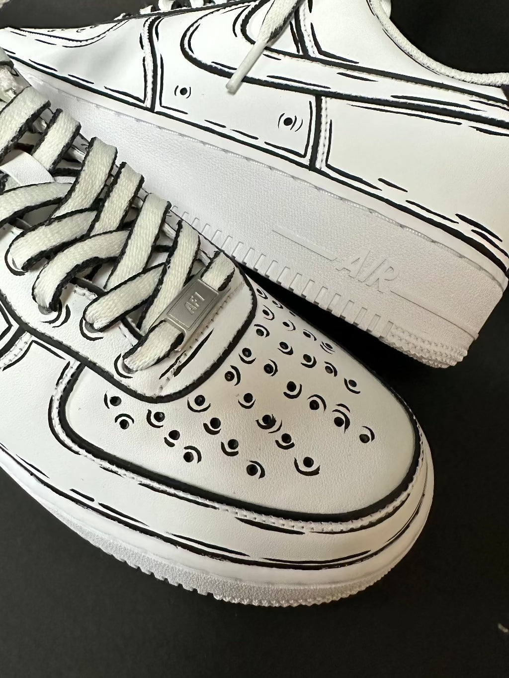 Close-up of white streetwear sneakers with black cartoon detailing and perforated toe box.