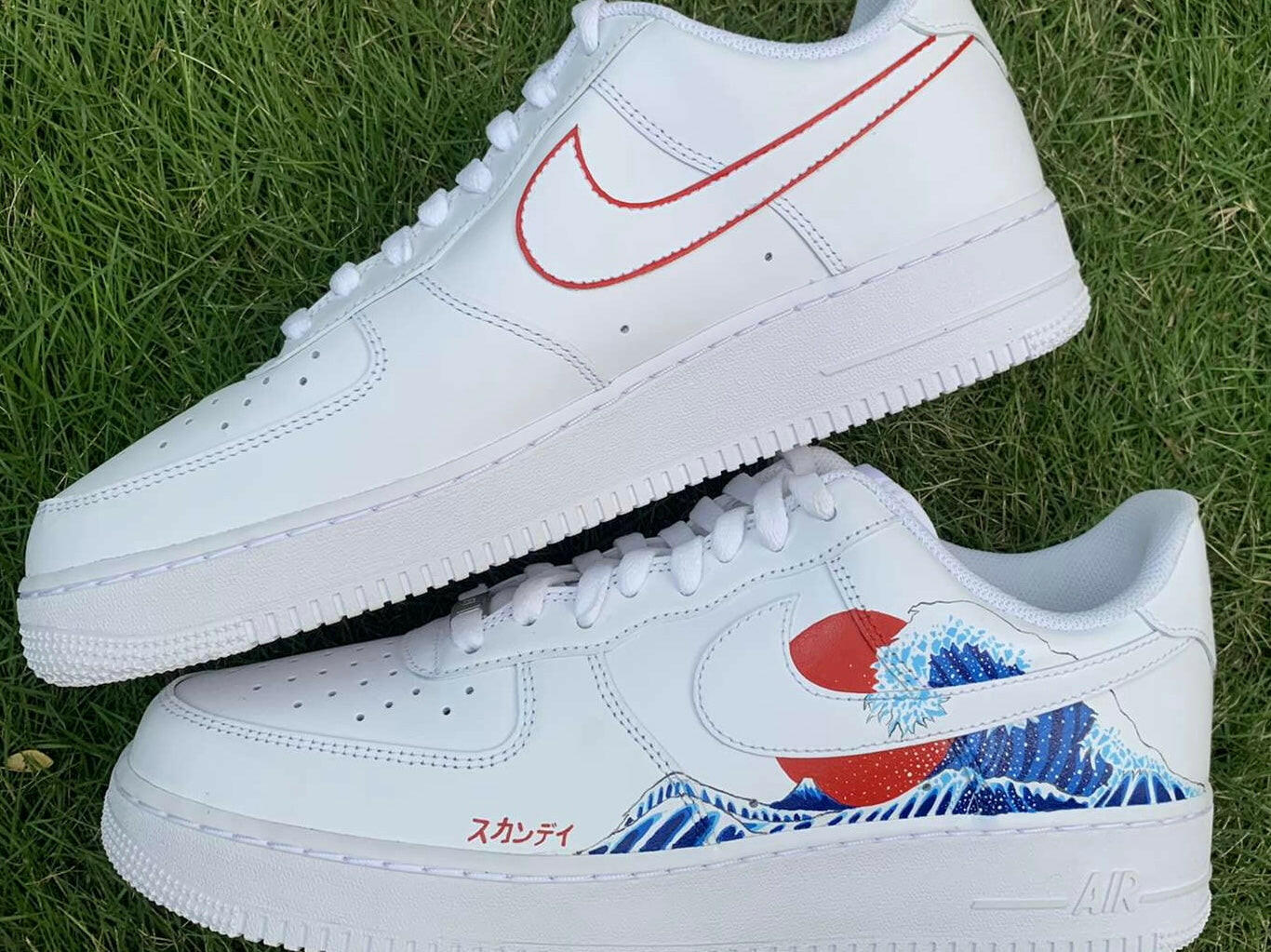 White streetwear sneakers on grass, one with red swoosh outline, one with wave and sun graphic