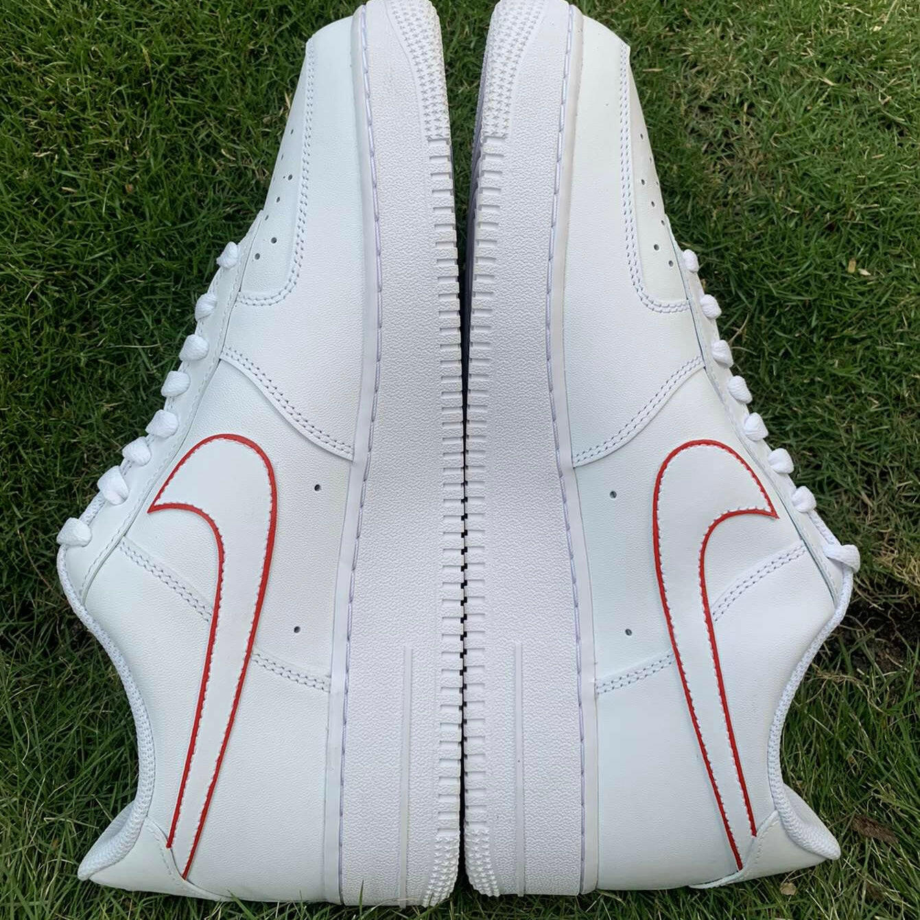 White leather sneakers with red outline swoosh logos side by side on green grass