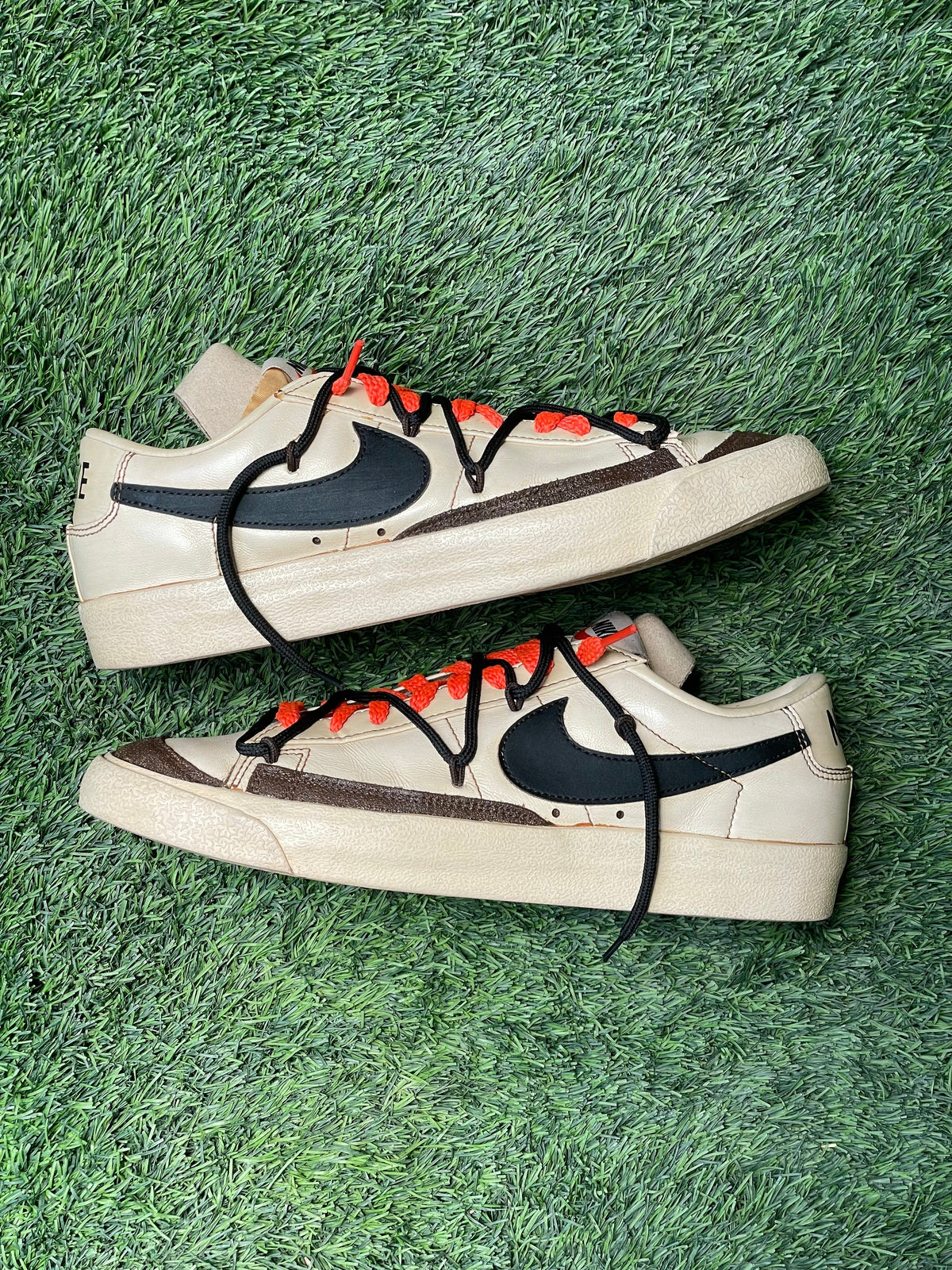 Beige low-top sneakers with black swoosh, orange laces, on green grass background