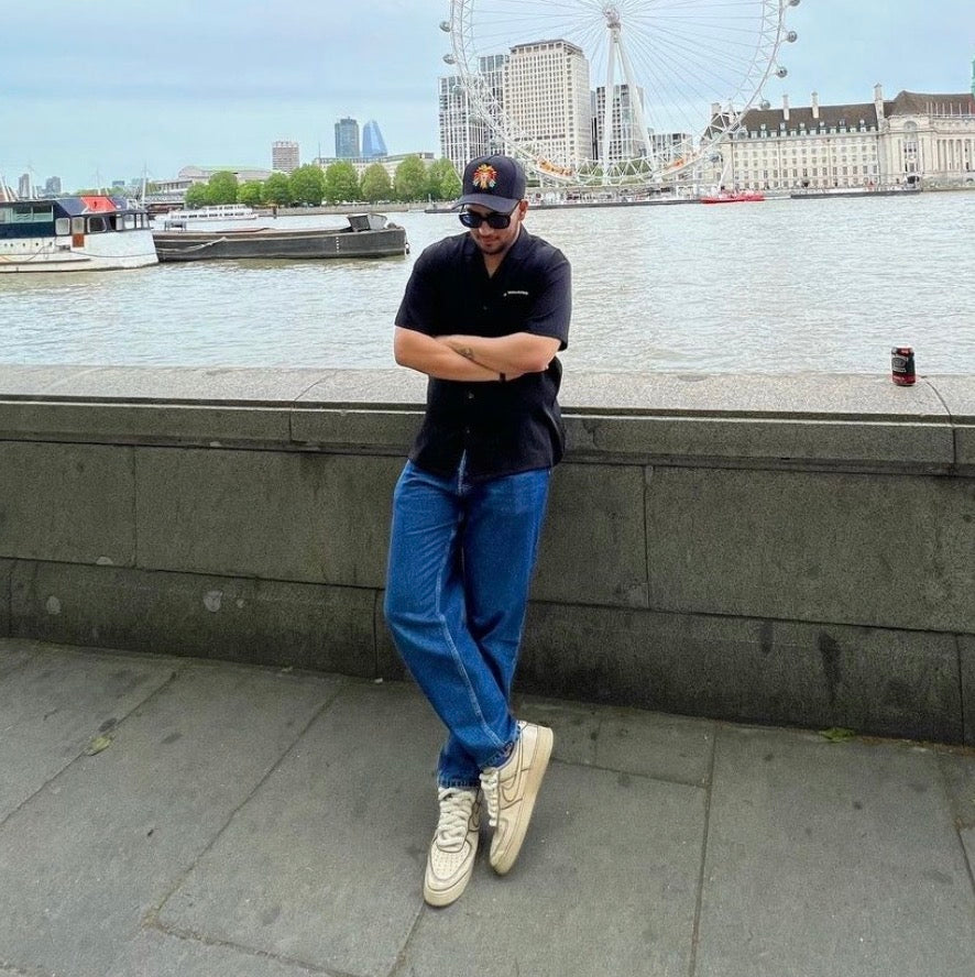 Man in streetwear outfit with sneakers and cap by river, London Eye in background