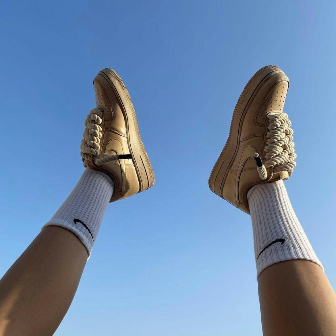 Beige streetwear sneakers with chunky laces worn with white socks against a clear blue sky.