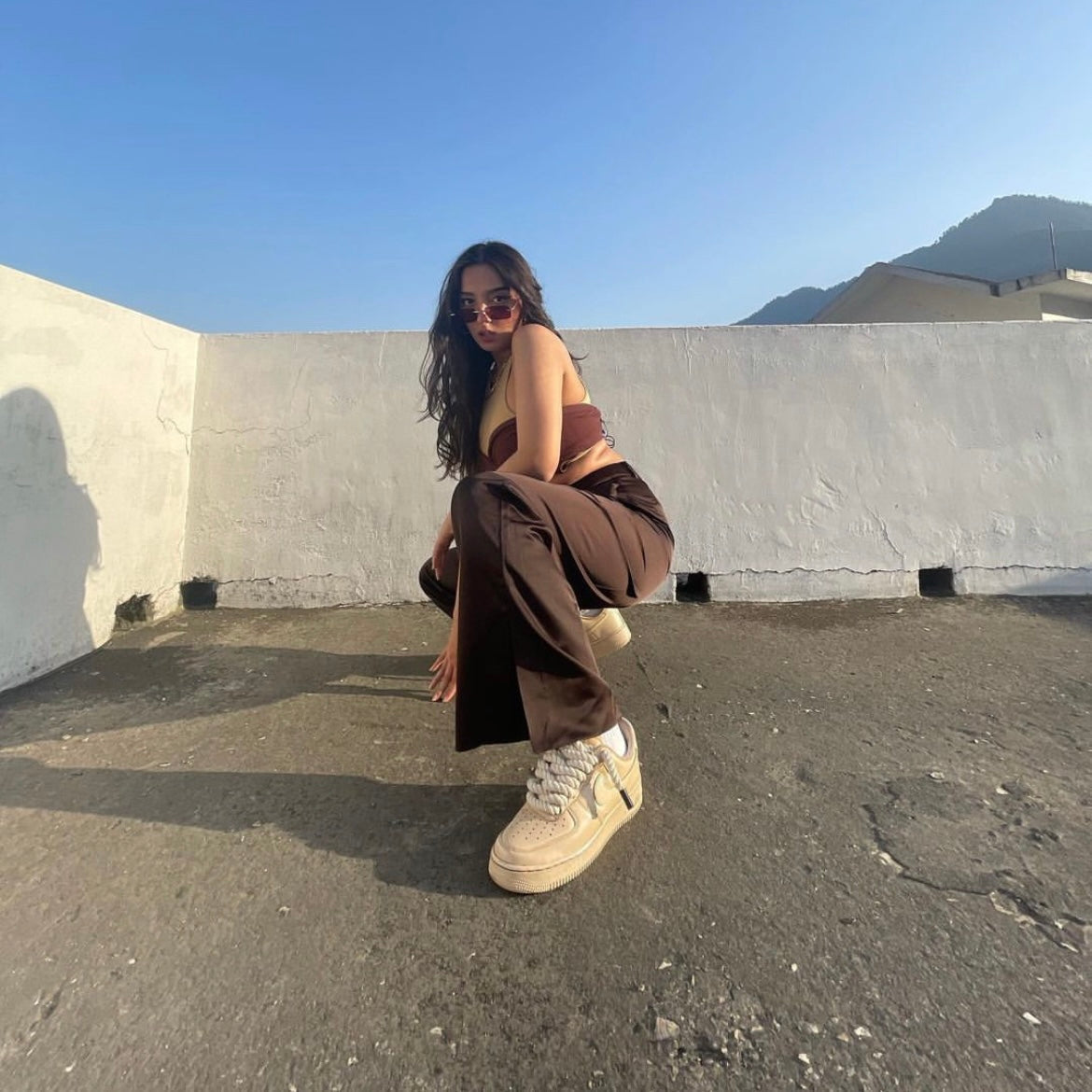 Woman in brown streetwear outfit and sunglasses squatting on rooftop in beige designer sneakers