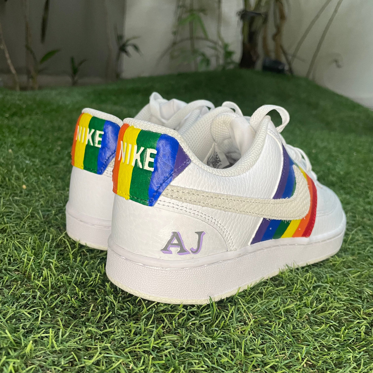 White Nike sneakers with rainbow stripes and custom 'AJ' detail on artificial grass