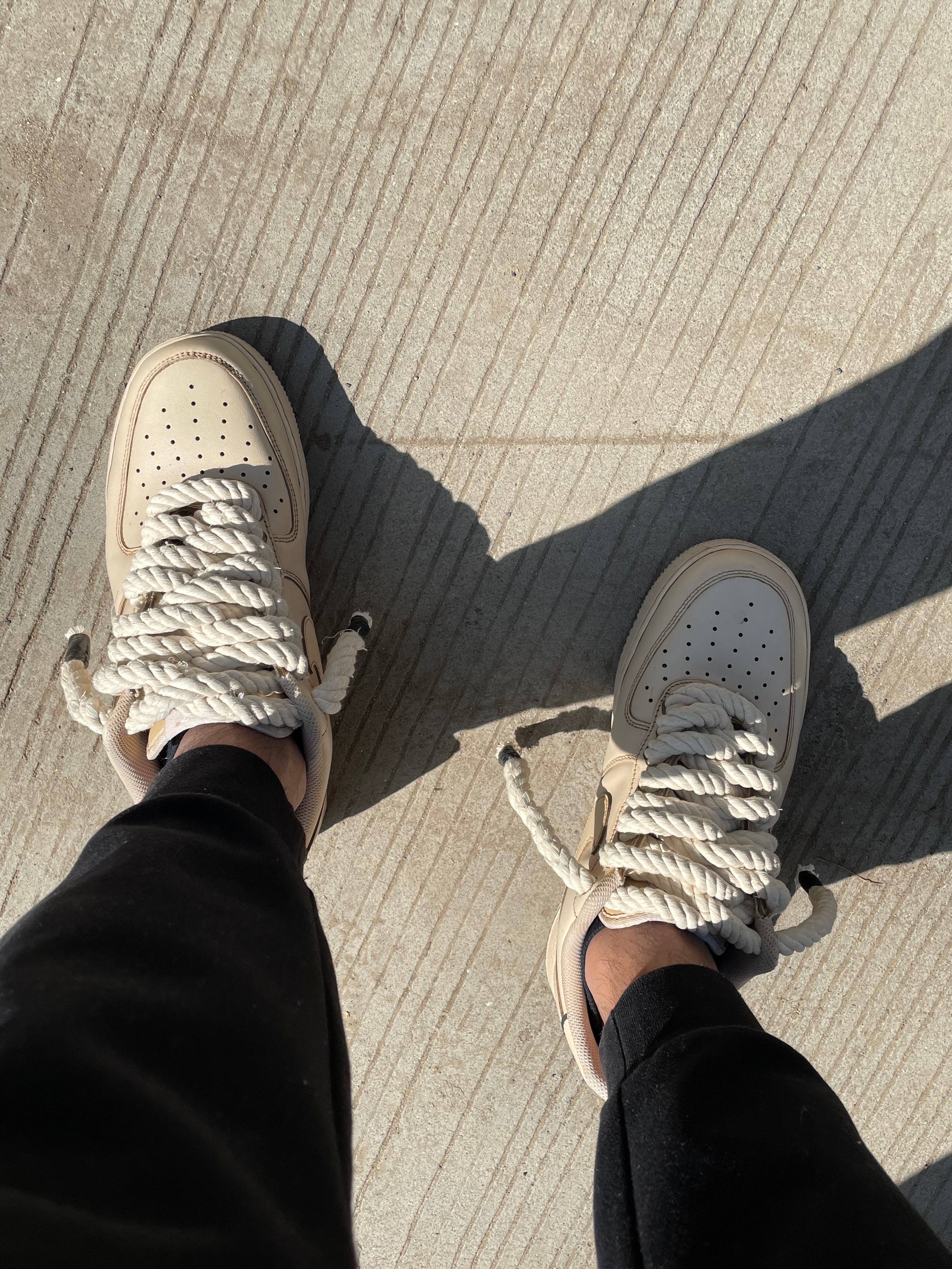 Beige streetwear sneakers with thick rope laces, worn outdoors with black pants