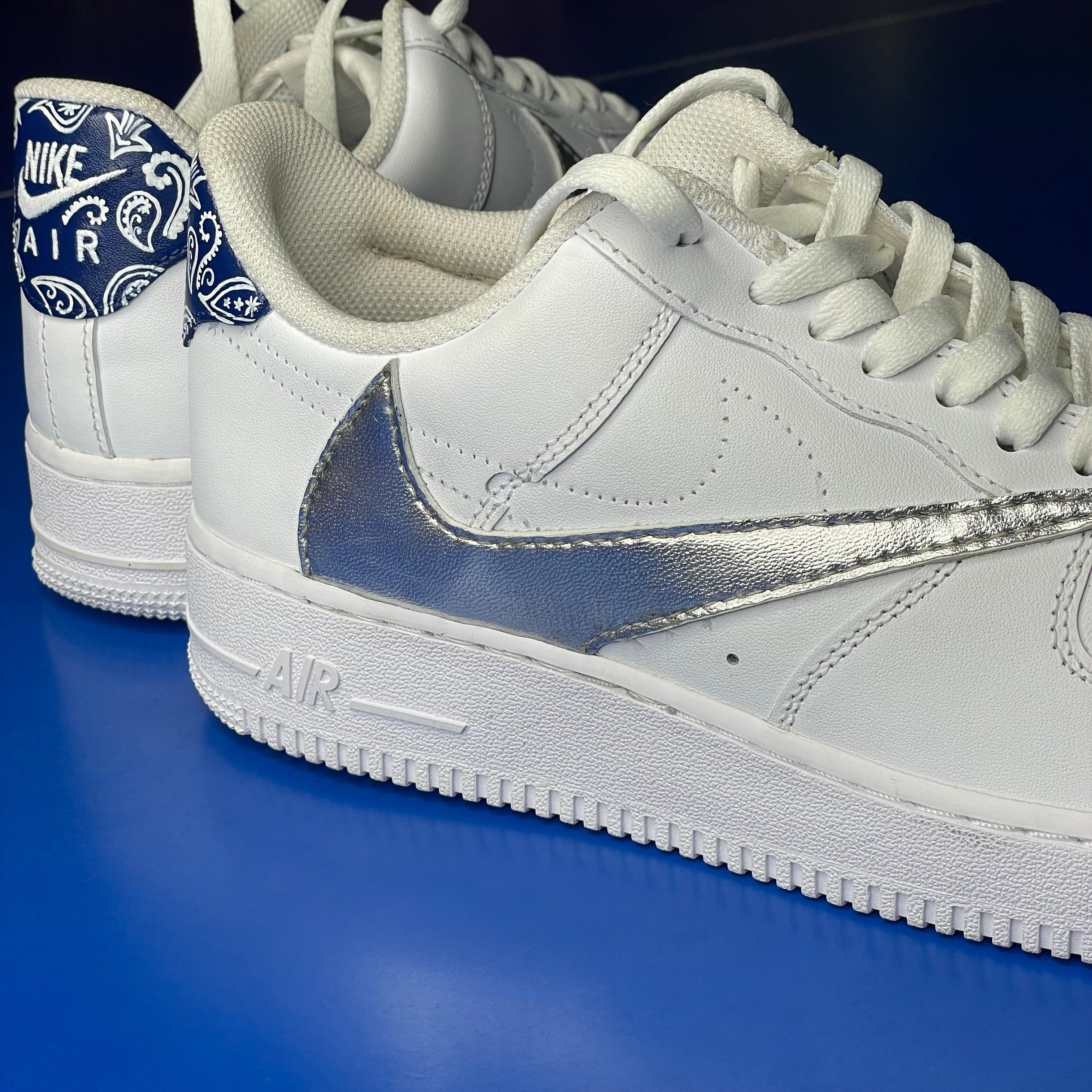 White Nike Air Force 1 sneakers with metallic silver swoosh and blue paisley heel detail on blue background