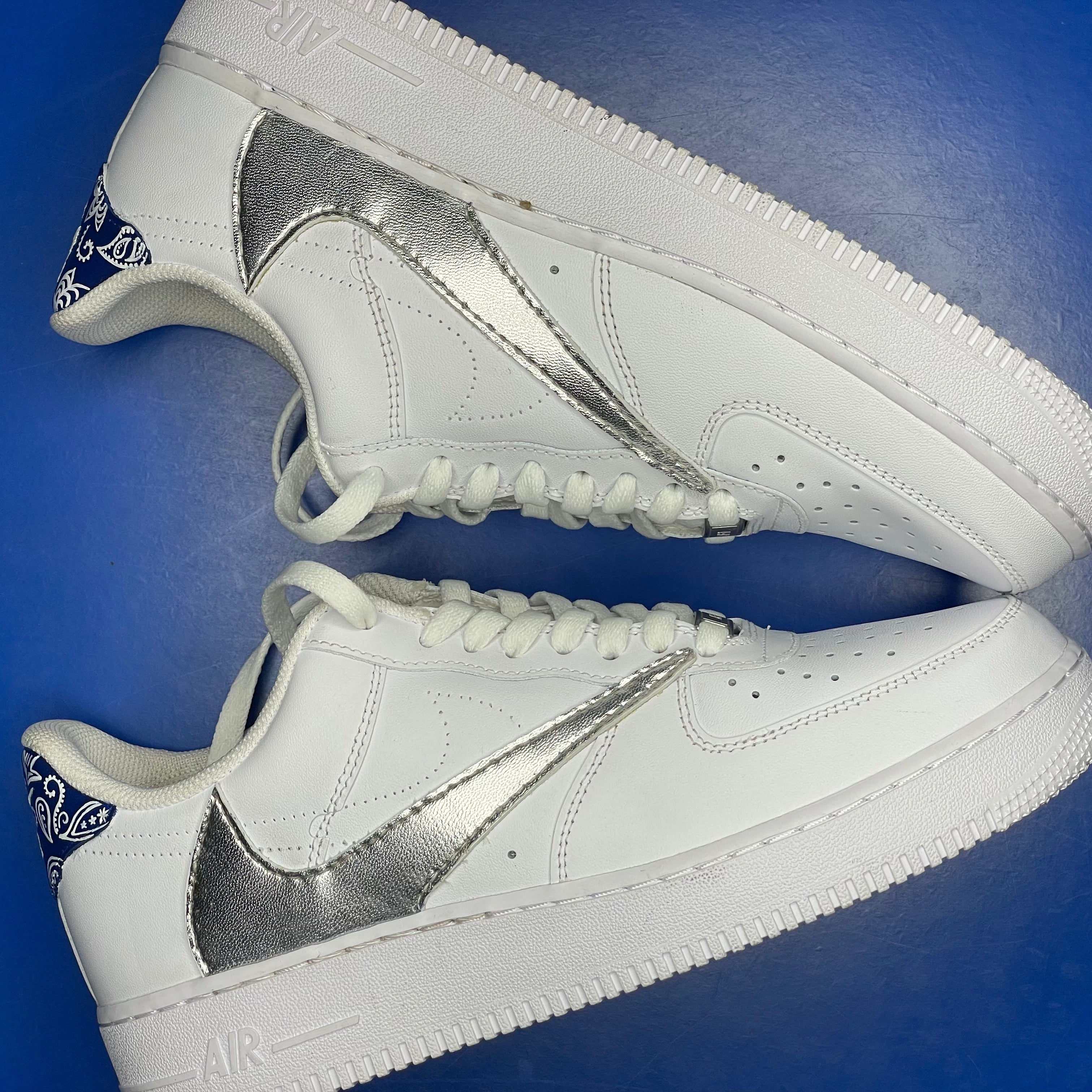 White streetwear sneakers with metallic silver swooshes and blue paisley heel detail on a blue background.