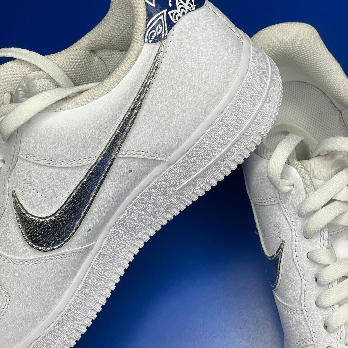 White leather sneakers with metallic silver swoosh and paisley heel detail on blue background
