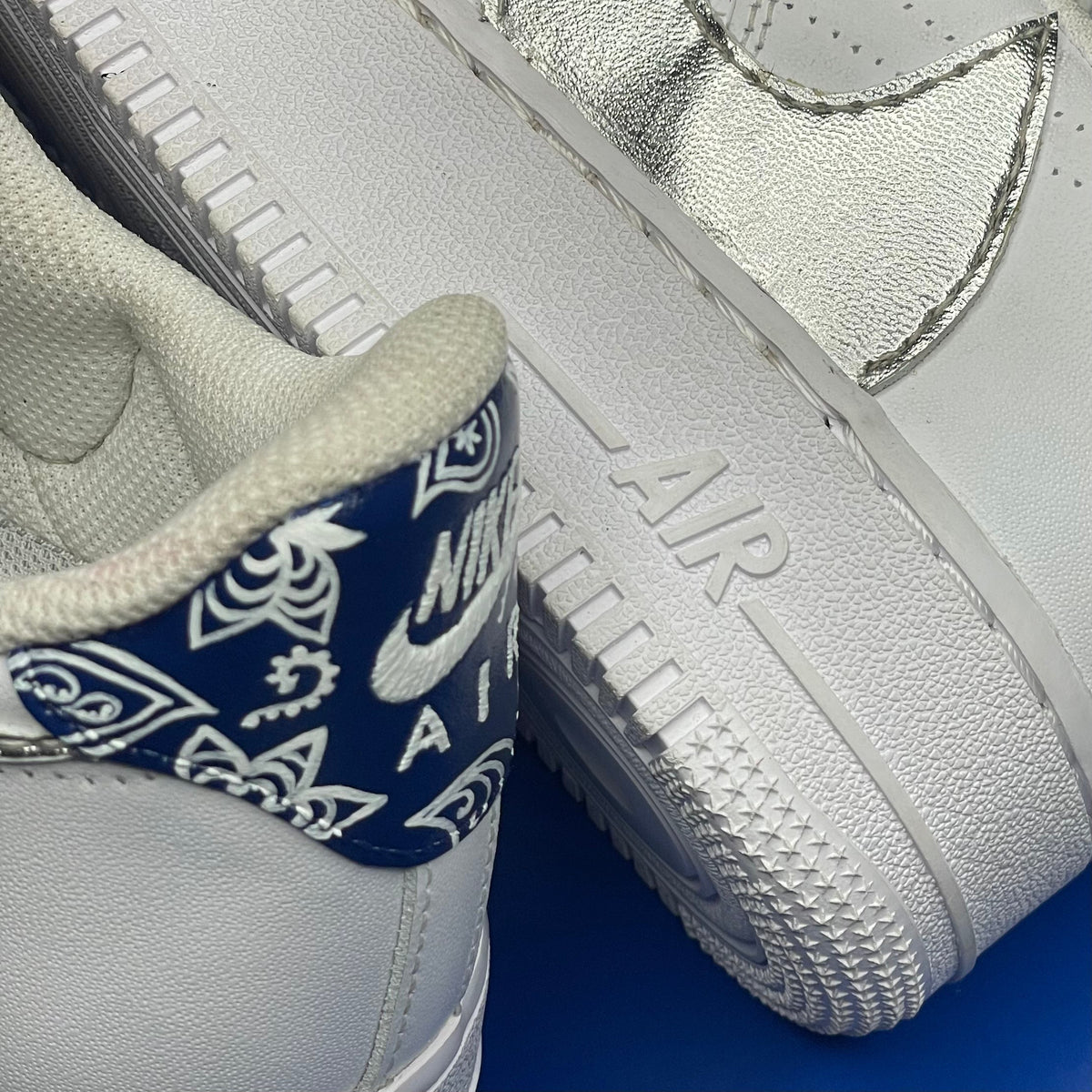White Nike Air Force 1 sneakers with metallic silver Swoosh and blue paisley heel tab on a blue background