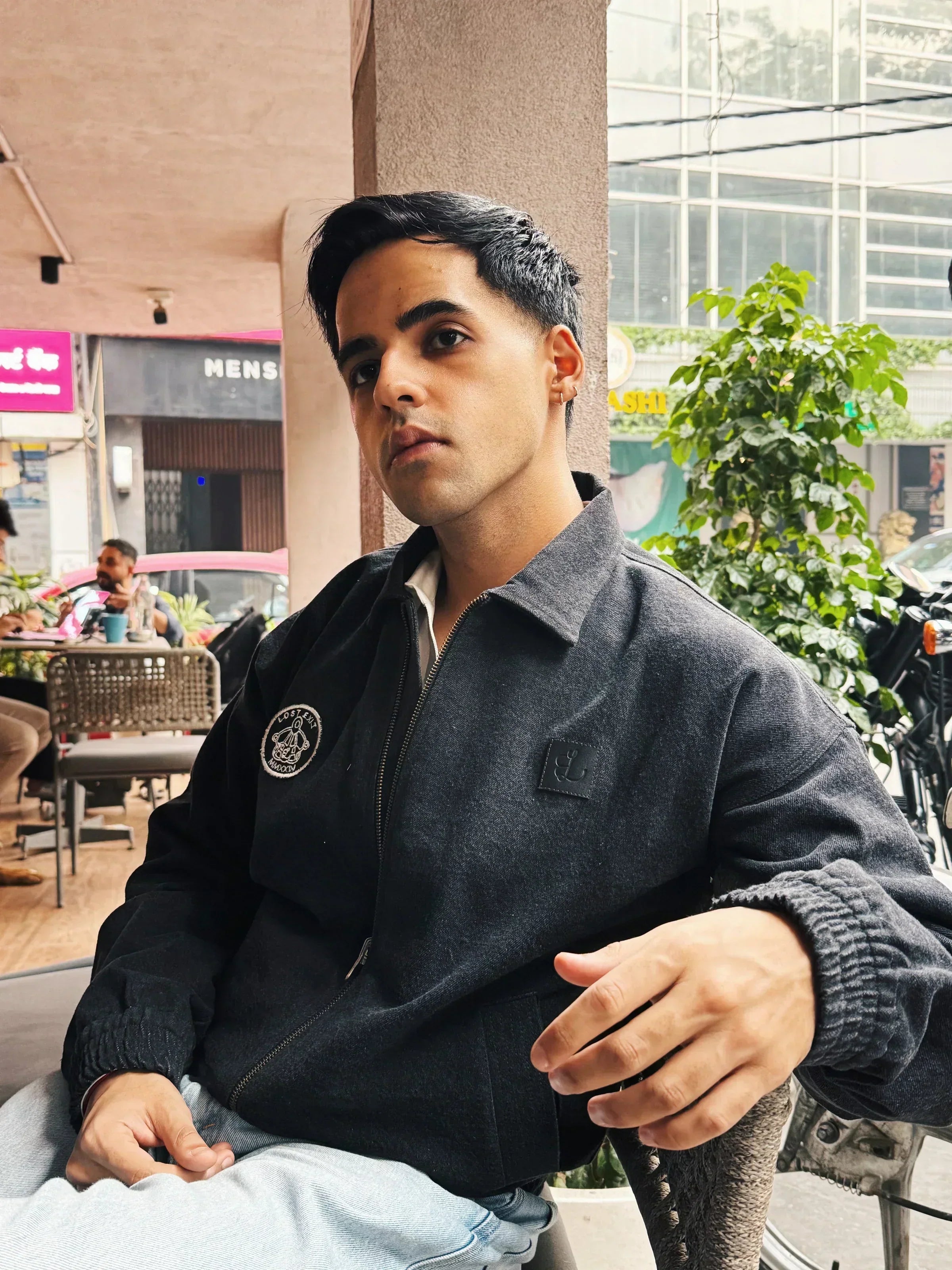 Young man in black jacket sitting outdoors at a café with plants and urban background
