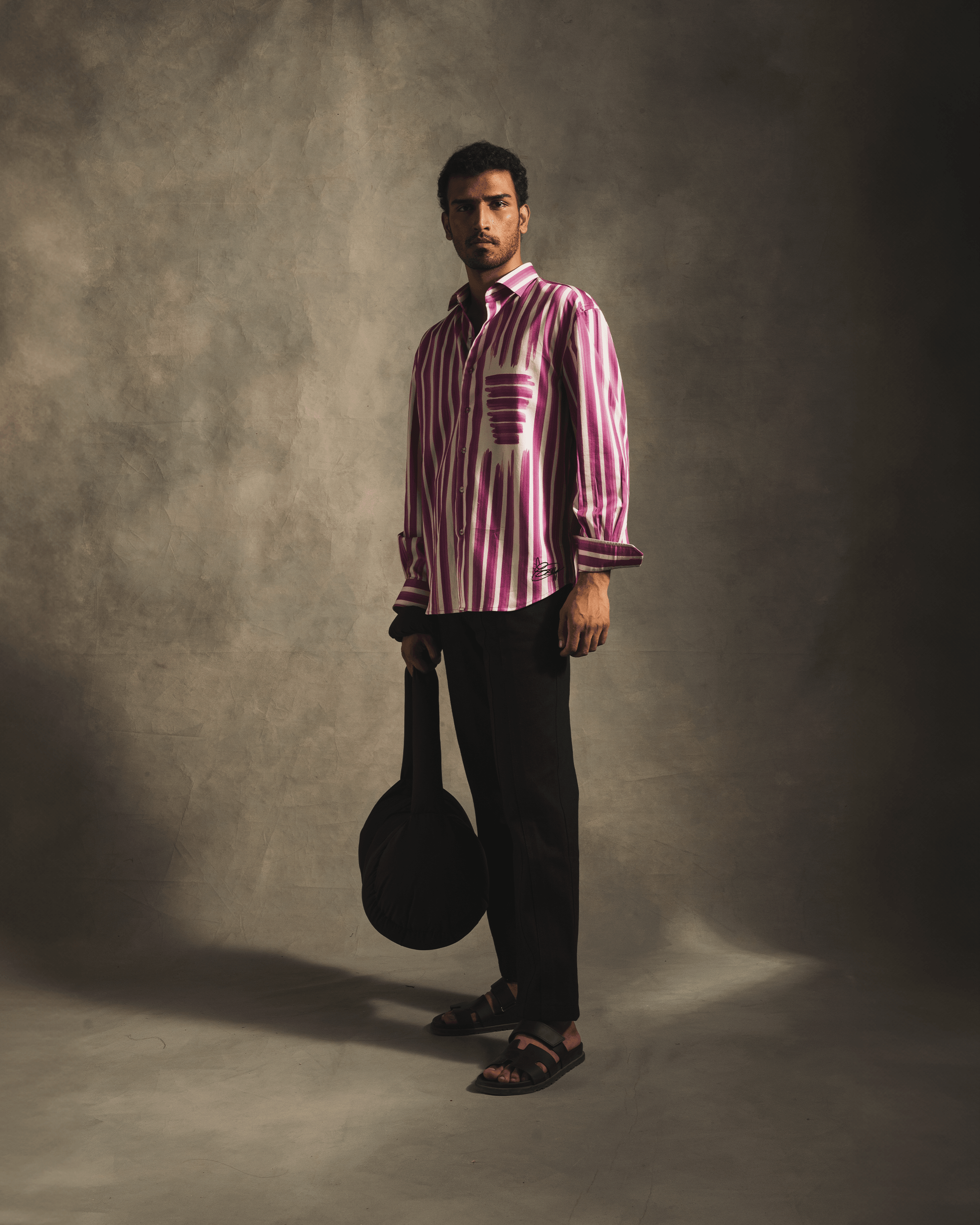 Man in a pink and white striped button-up shirt, black pants, black sandals, holding a round black bag in a studio setting.