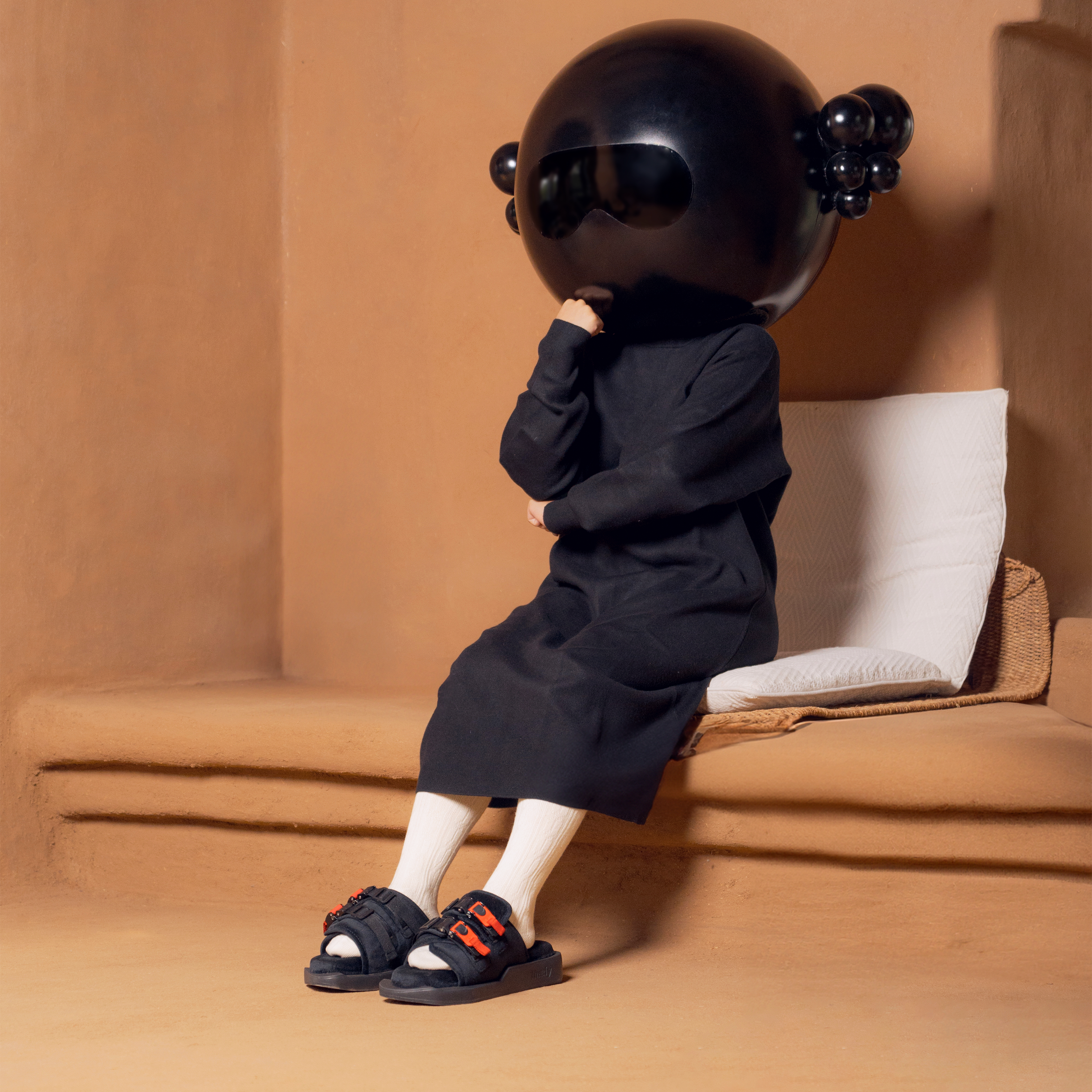 Person in black dress and unique black helmet sitting on cushion, wearing black sandals with red accents, in a minimal earthy-toned room.