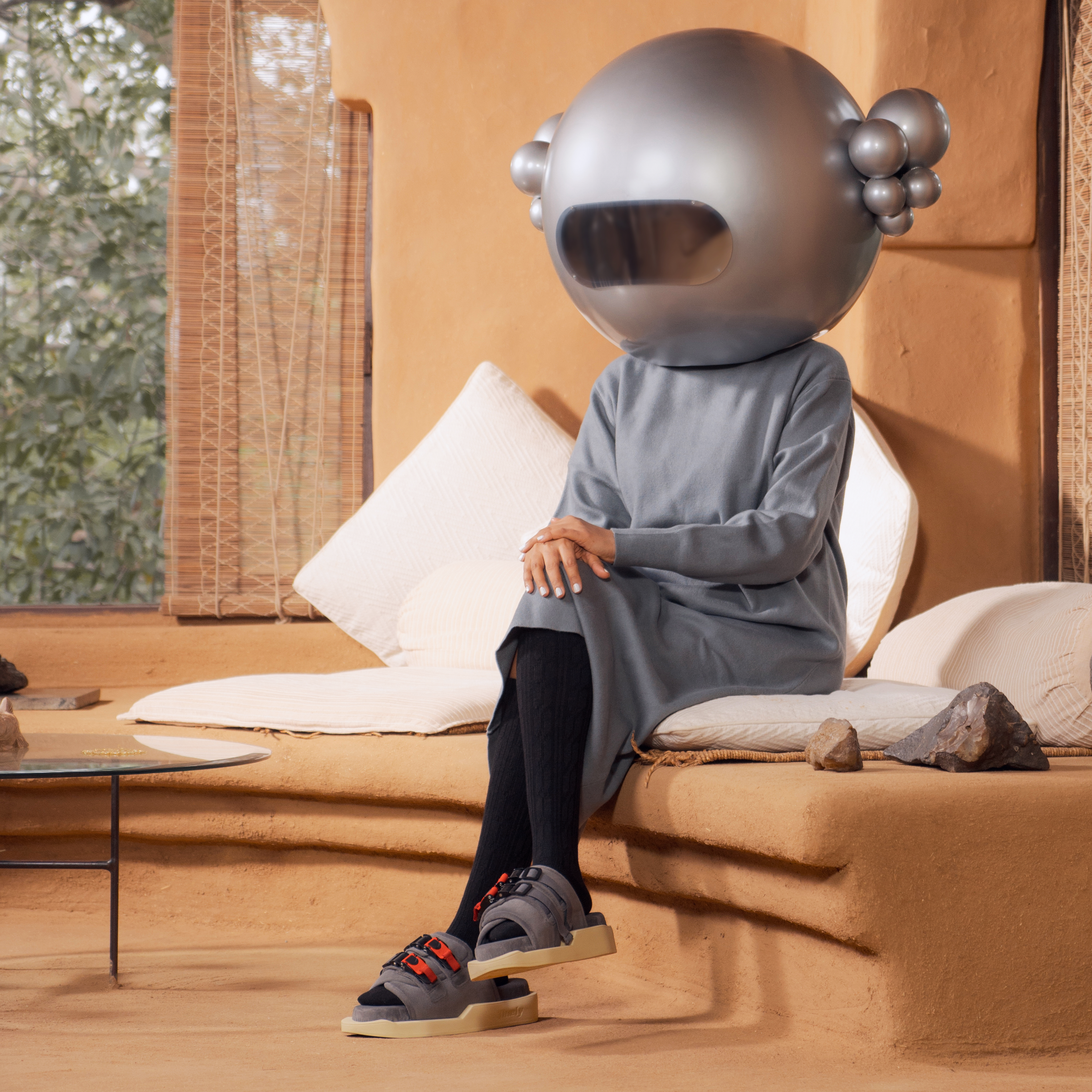 Person in futuristic silver helmet and grey streetwear, sandals with red straps, sitting indoors