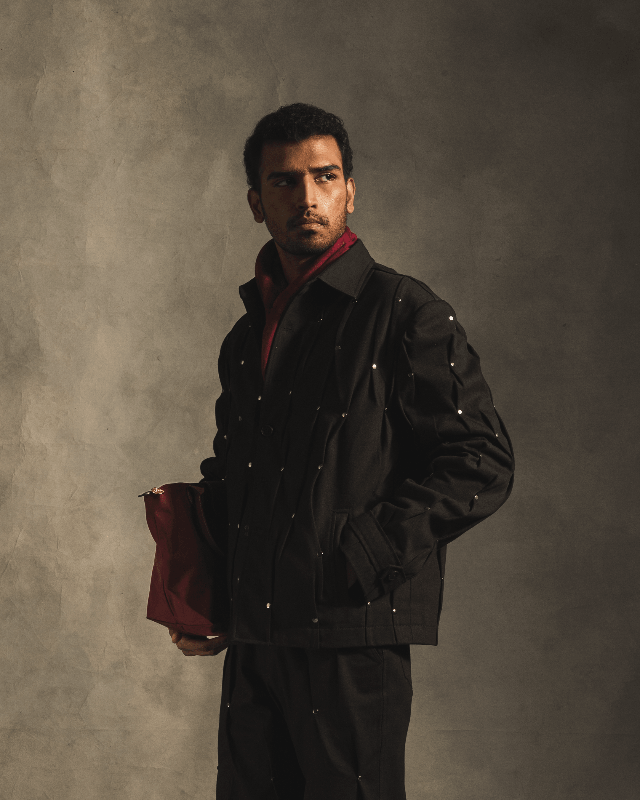 Man modeling a black rivet jacket and pants set, holding a red bag, streetwear fashion