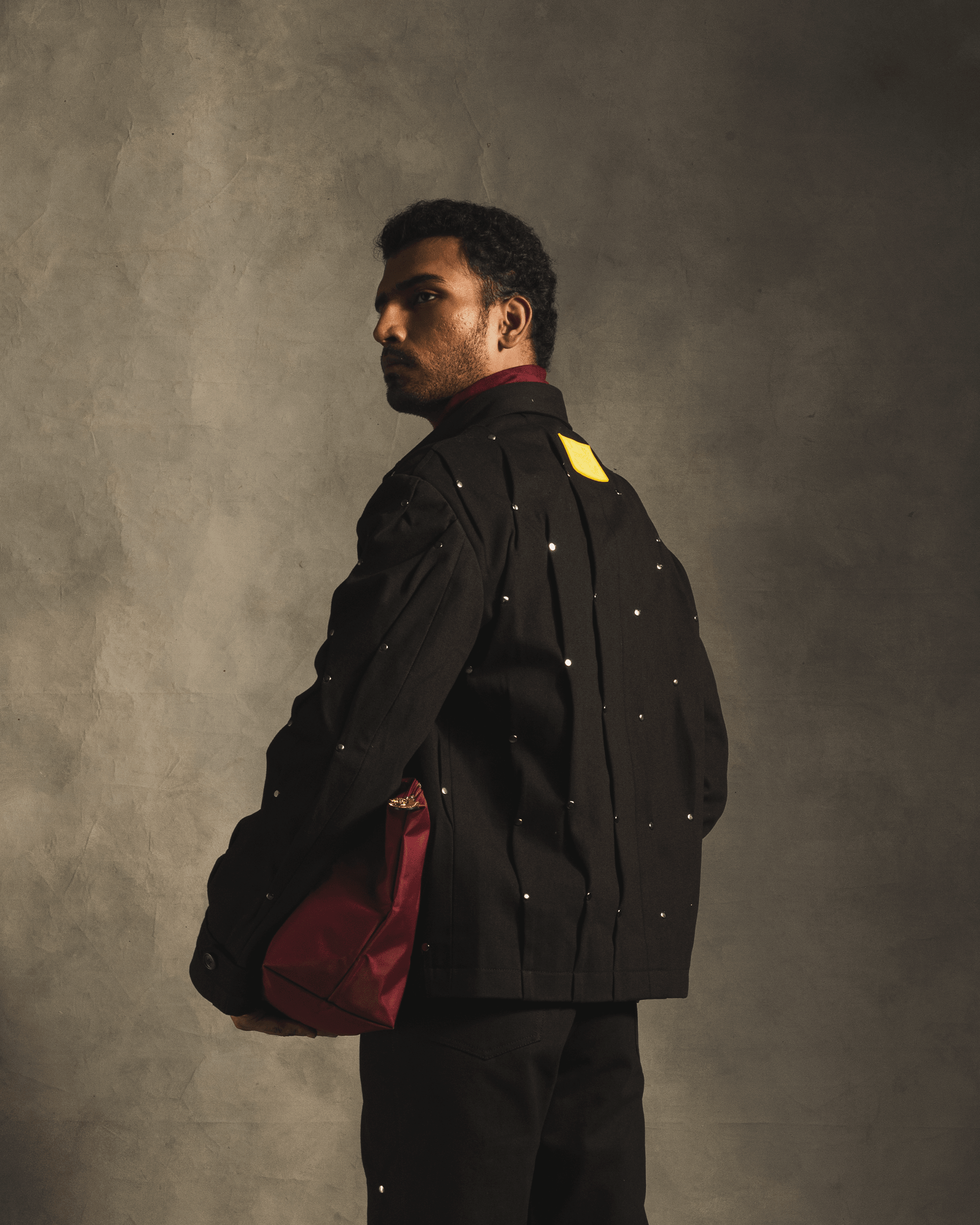 Man in a black rivet jacket with yellow label, holding a red bag, streetwear fashion