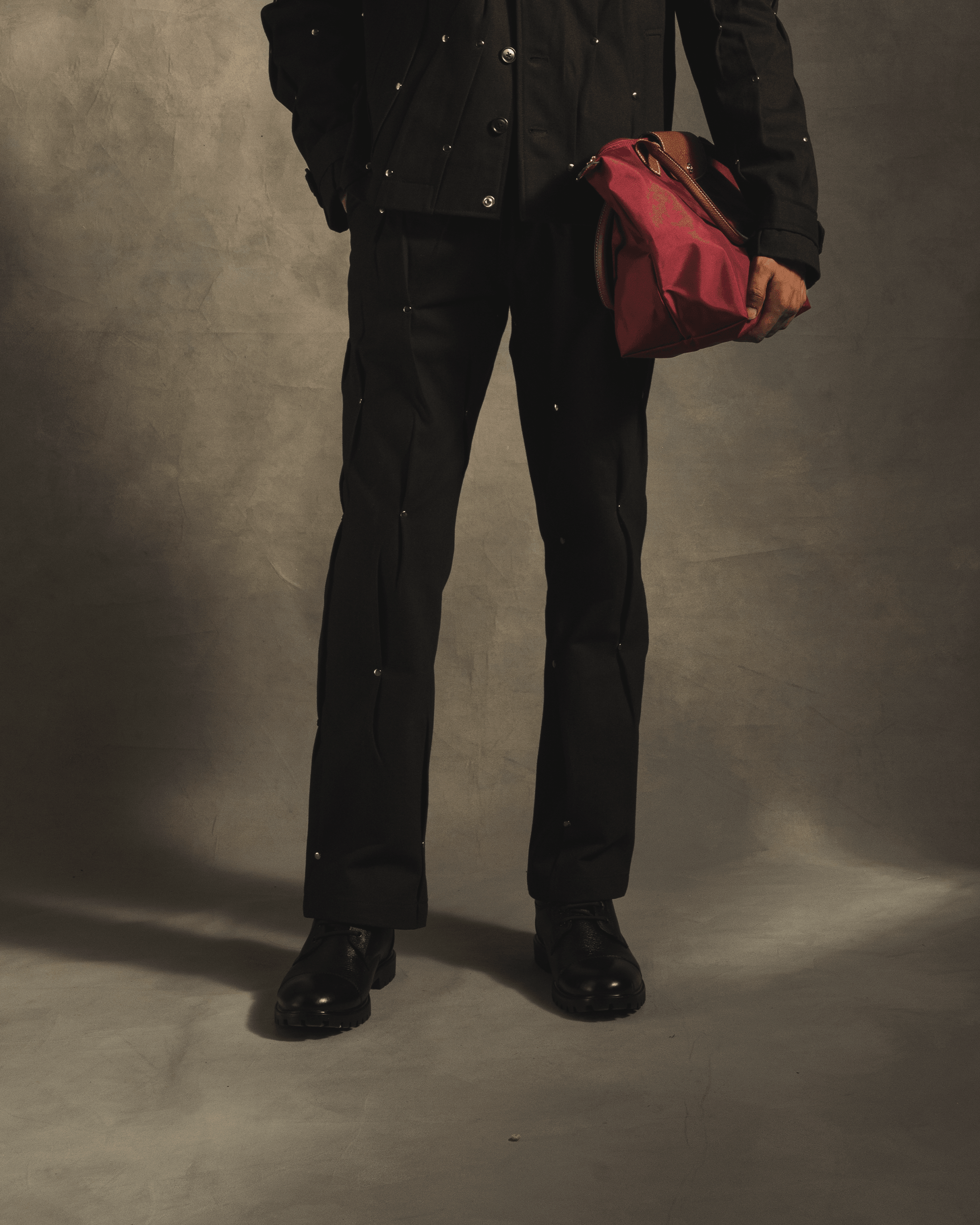 Black rivet pants paired with matching jacket, black shoes, and a red duffel bag in studio
