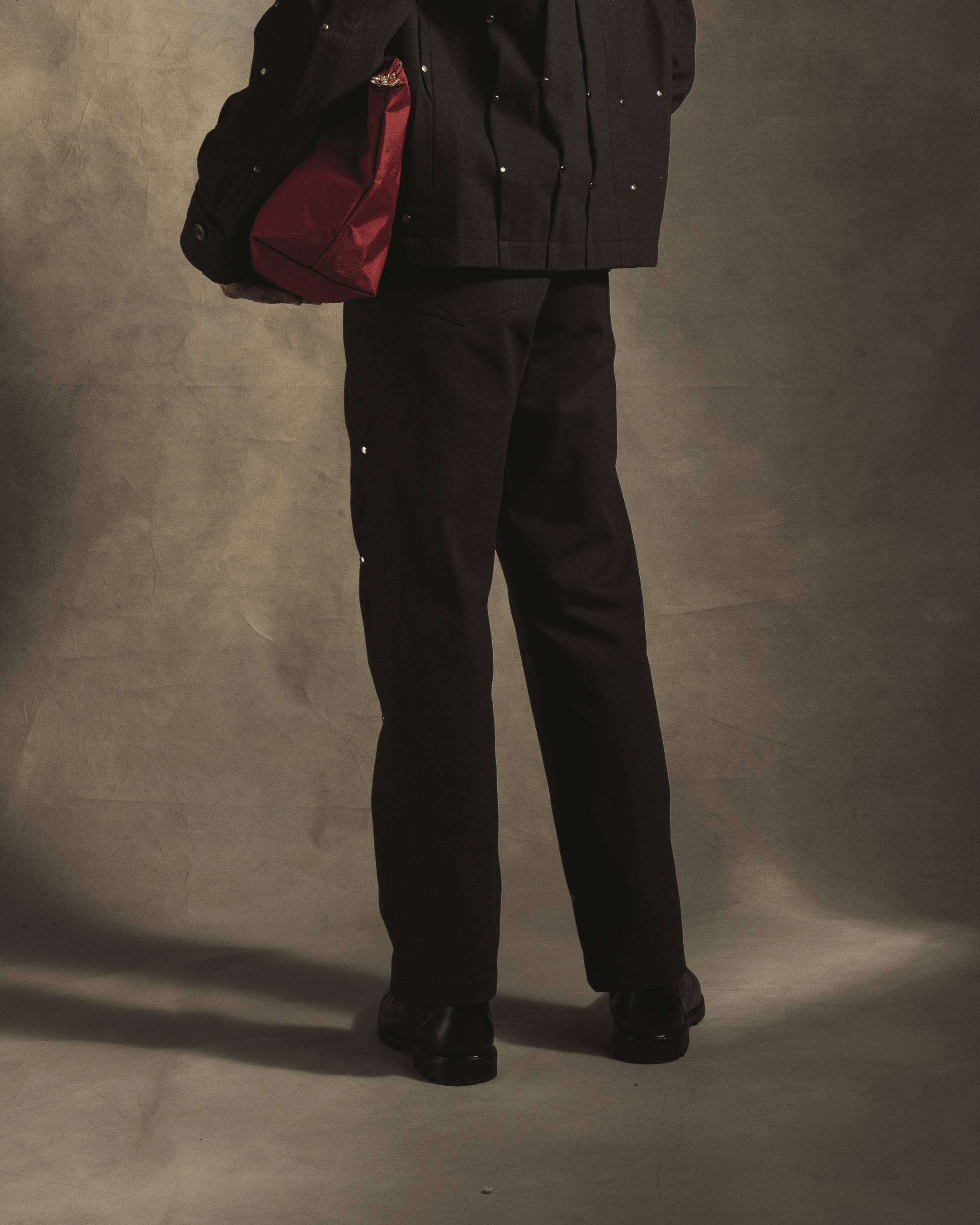 Back view of black streetwear pants with metal rivet details, person holding red bag