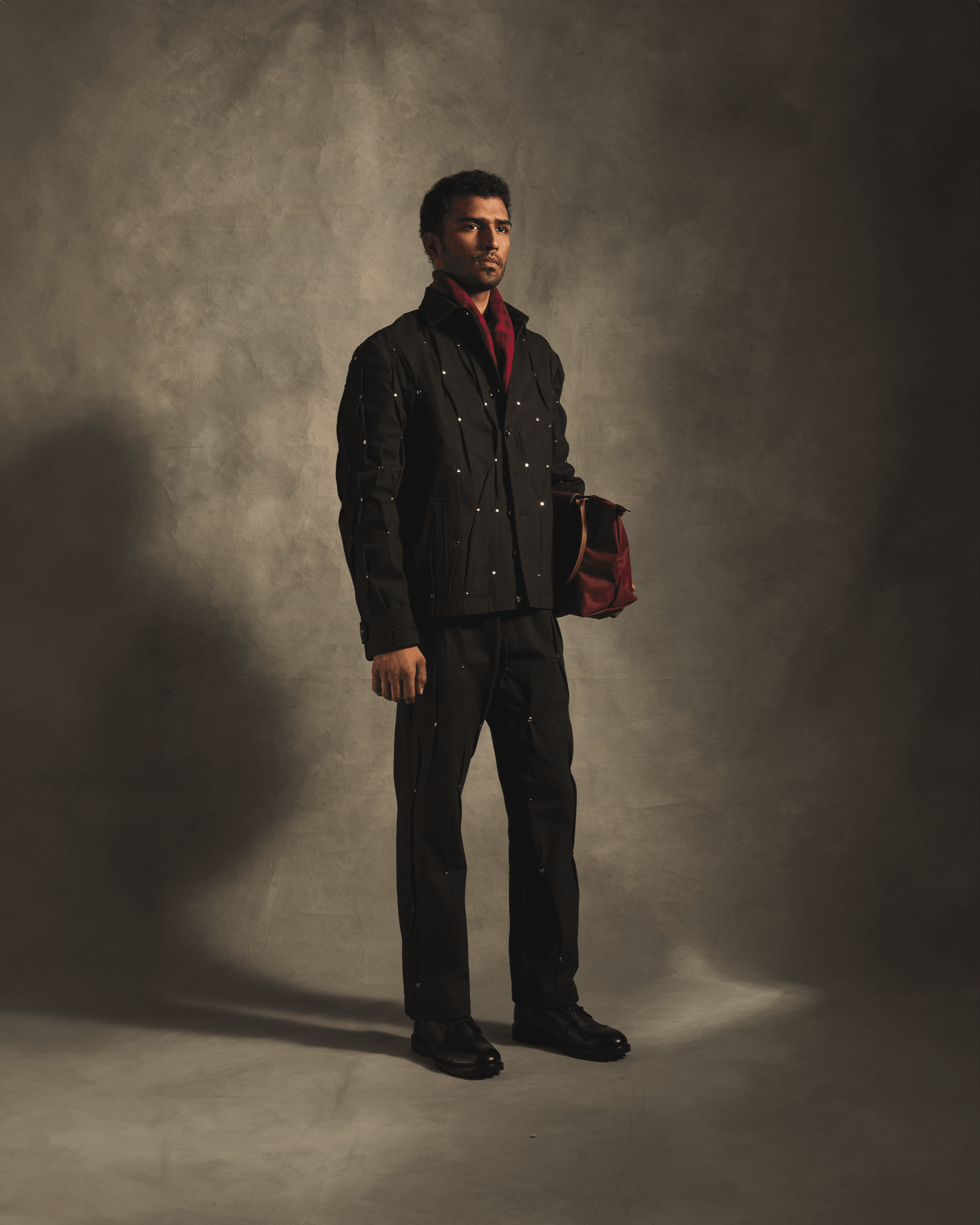 Man in black streetwear suit with silver rivet details, holding a red bag, studio background