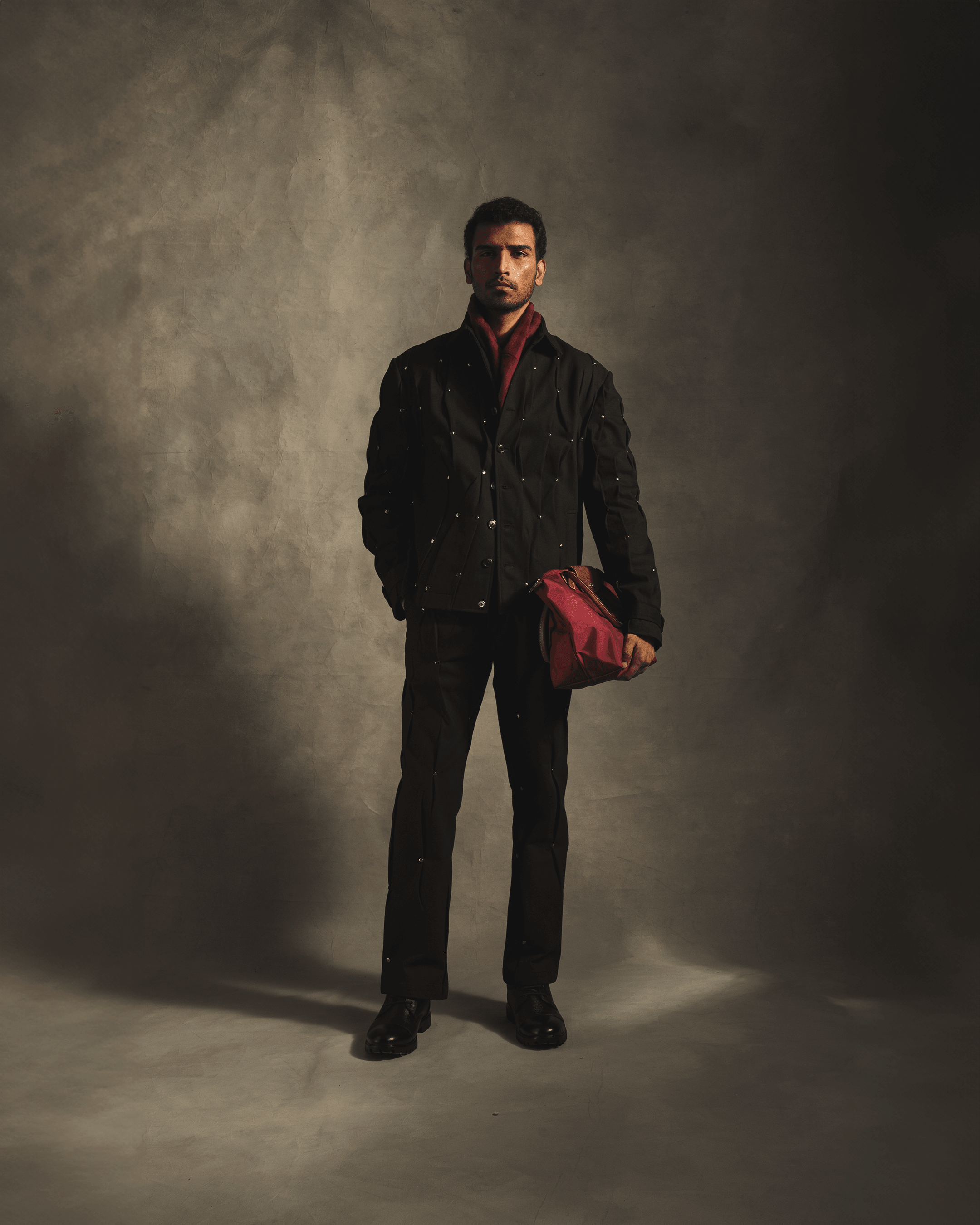 Man wearing black streetwear jacket and pants with metallic rivet details, holding a red bag