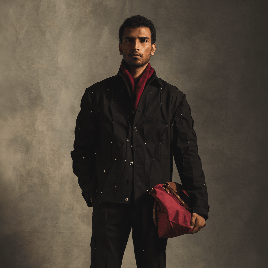 Man wearing black studded streetwear jacket and matching pants, holding red duffel bag