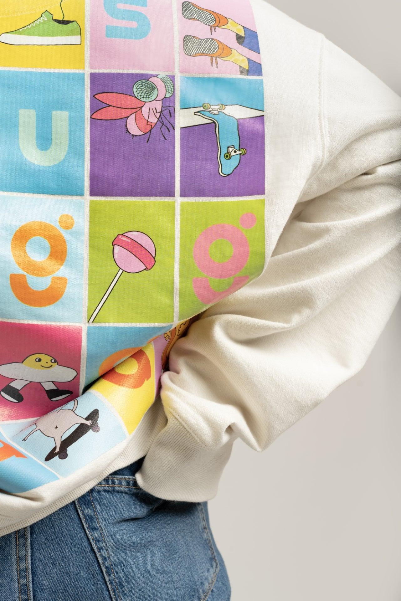Close-up of a cream streetwear sweatshirt with colorful cartoon patchwork graphic, worn with jeans.
