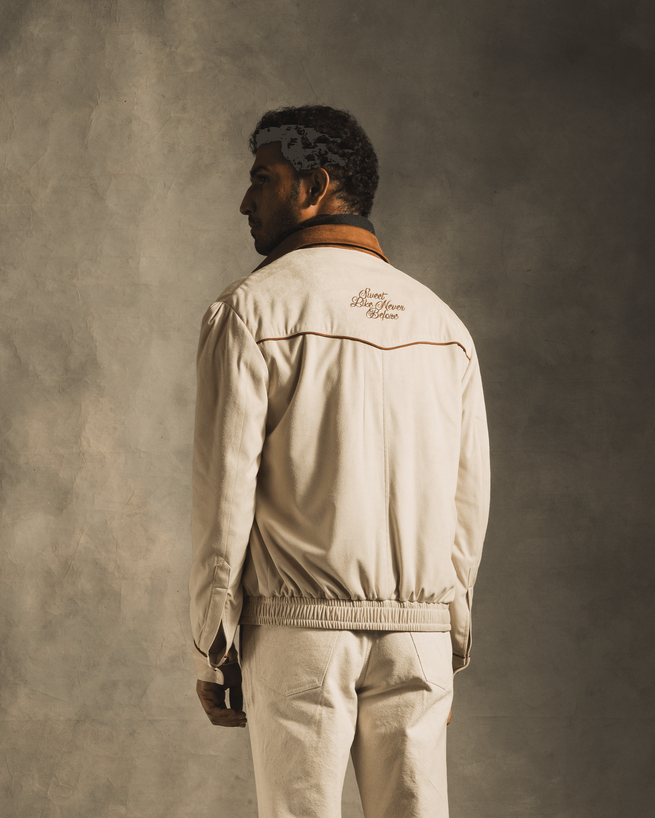 Man in beige suede jacket and matching pants, back view, embroidered text on jacket