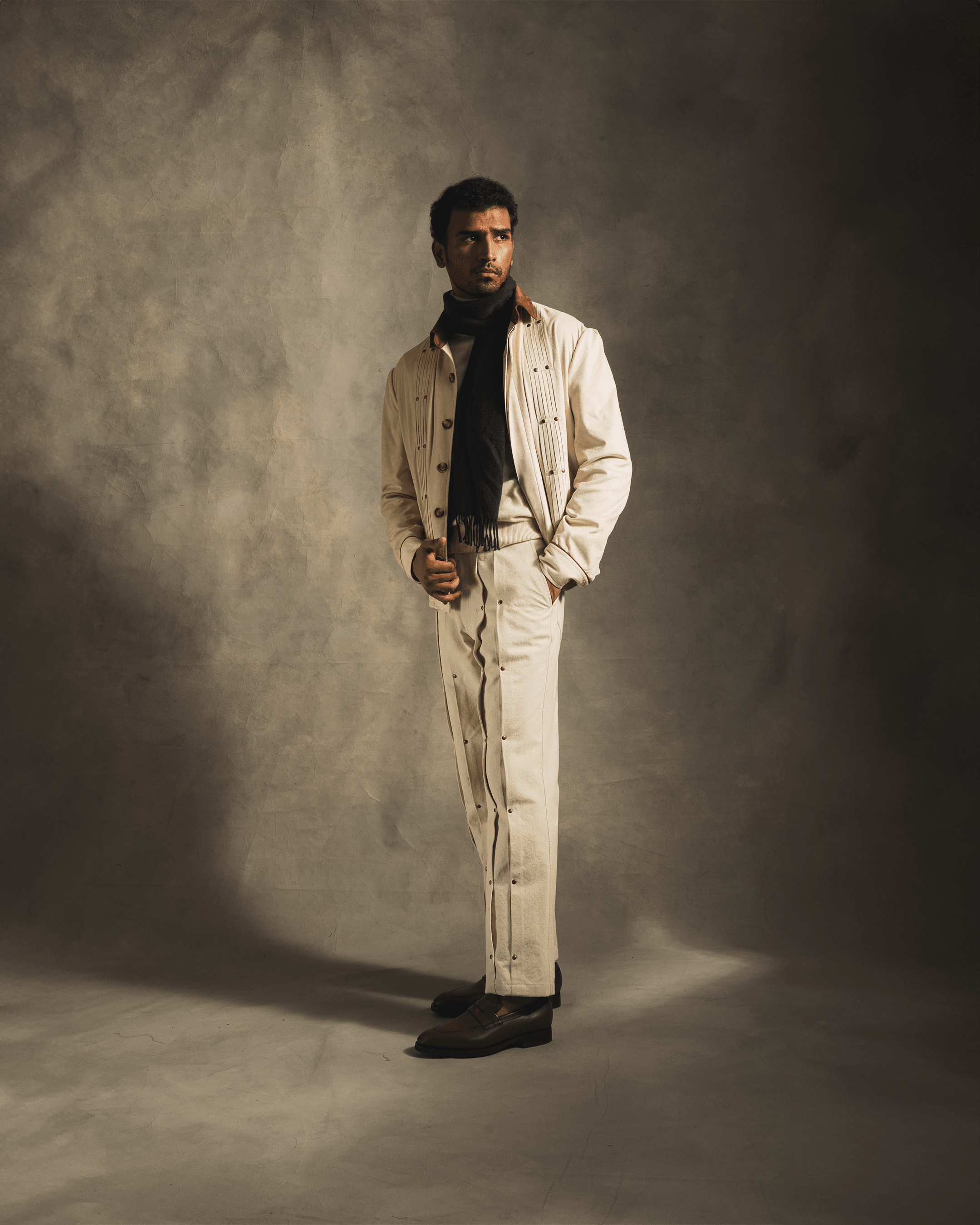 Man in beige suede jacket and matching pants with black scarf, posing in studio fashion shoot