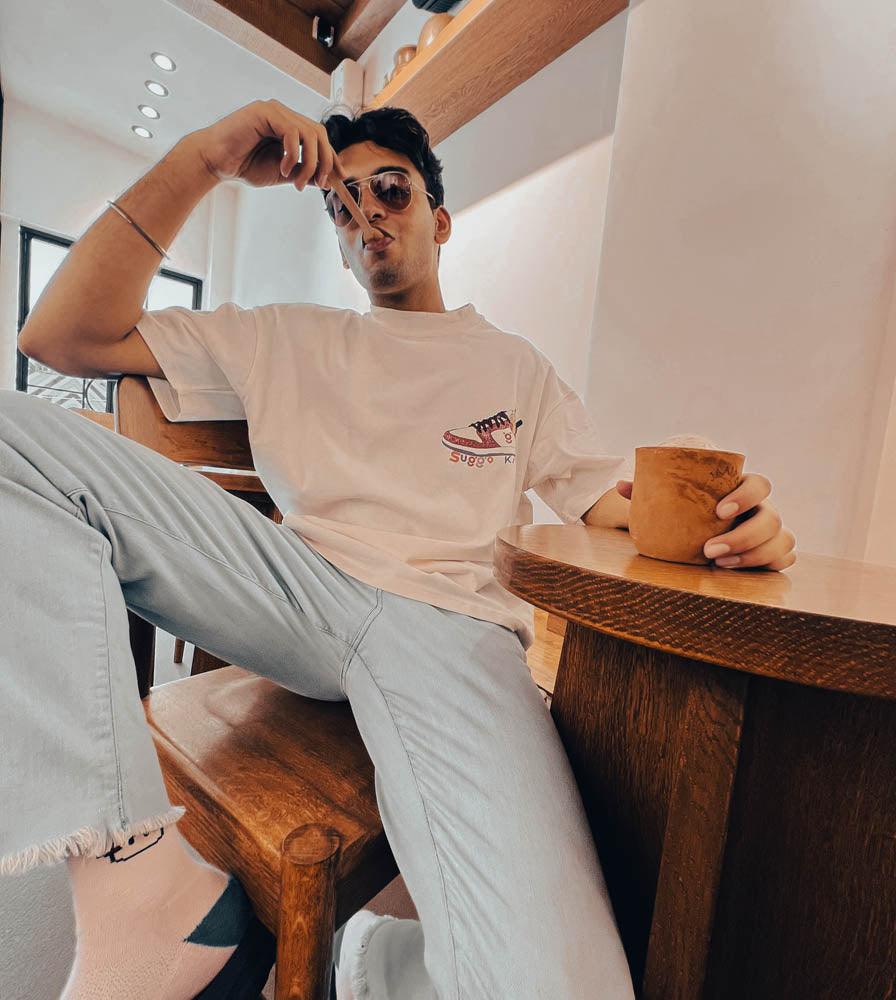 Man in casual streetwear with graphic sneaker t-shirt, light jeans, sunglasses, sitting in a cafe.