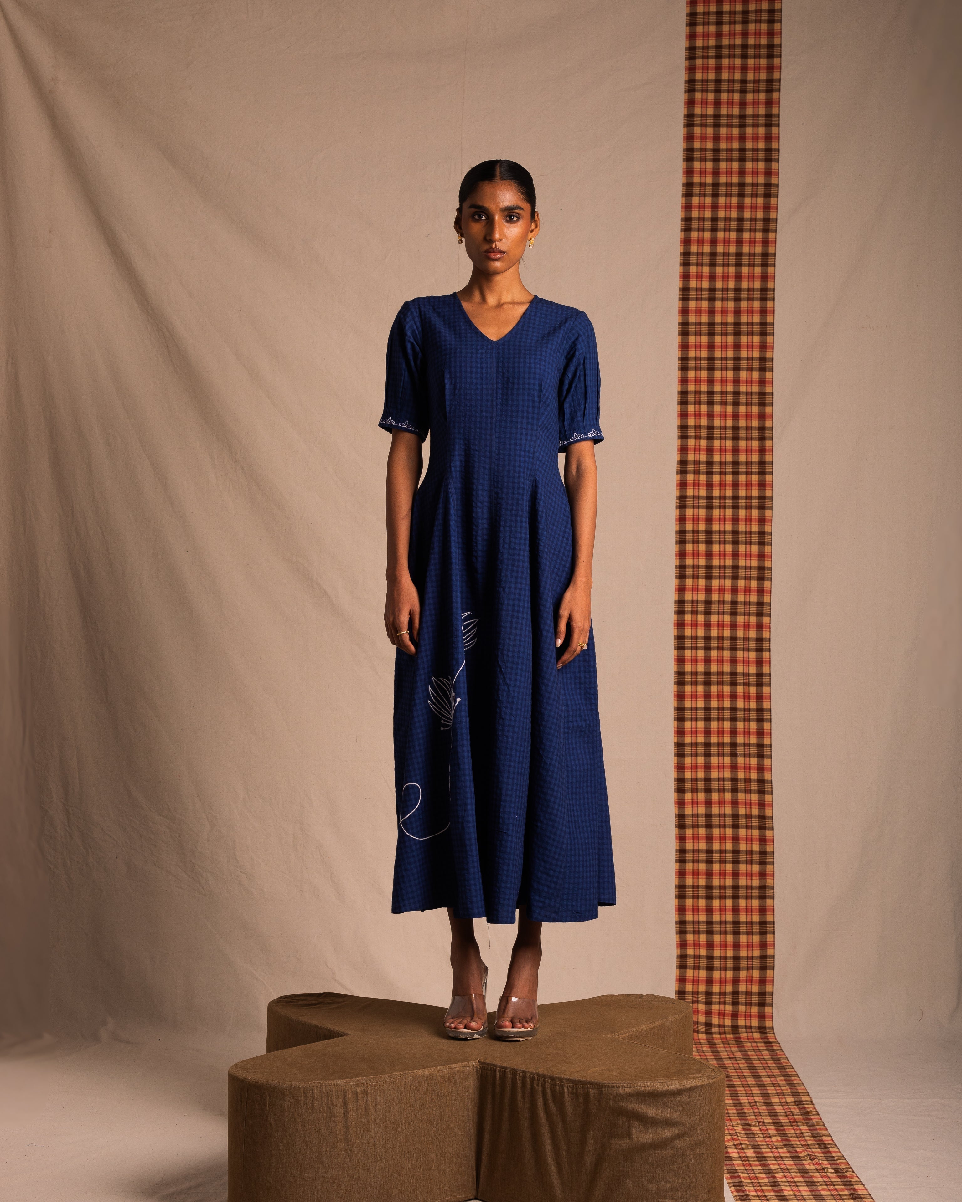 Woman in blue checkered midi dress with short sleeves and embroidery, standing on a brown platform.