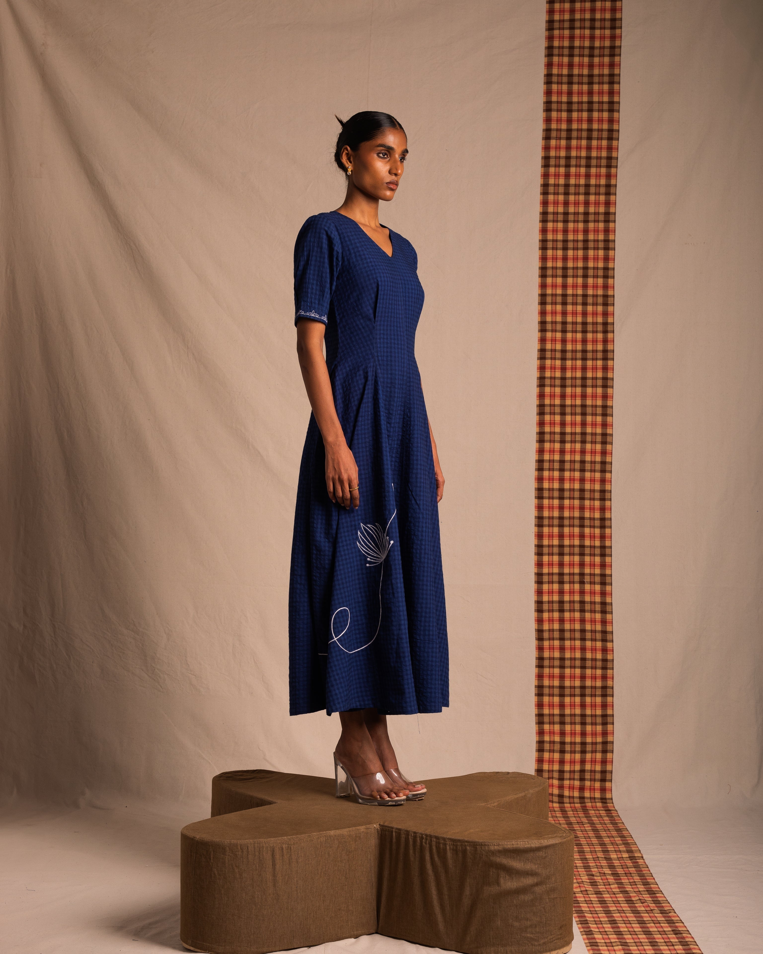 Woman modeling blue midi dress with V-neck and floral embroidery, standing on geometric platform