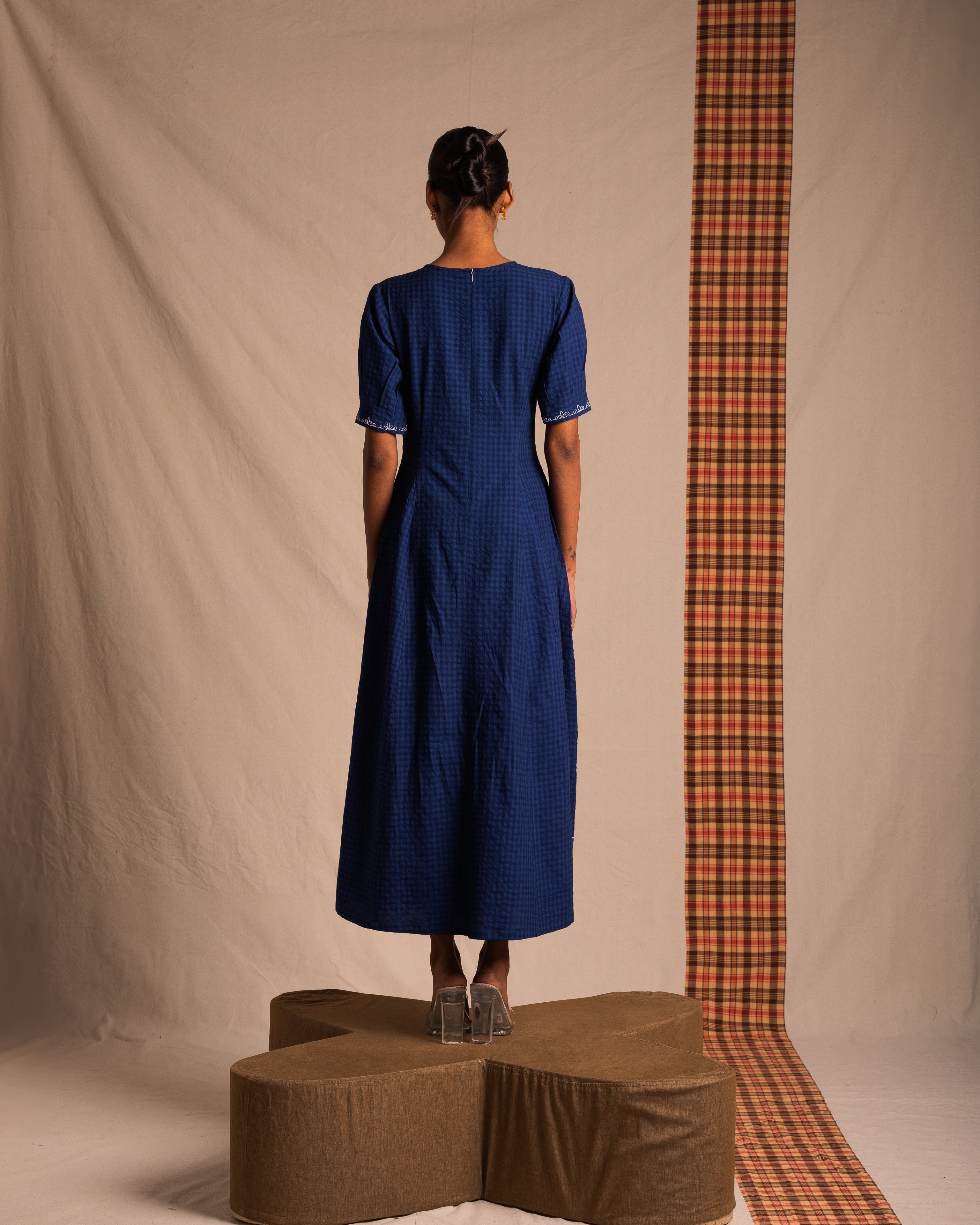 Woman in a blue checkered midi dress with short sleeves, standing on a platform, photographed from behind