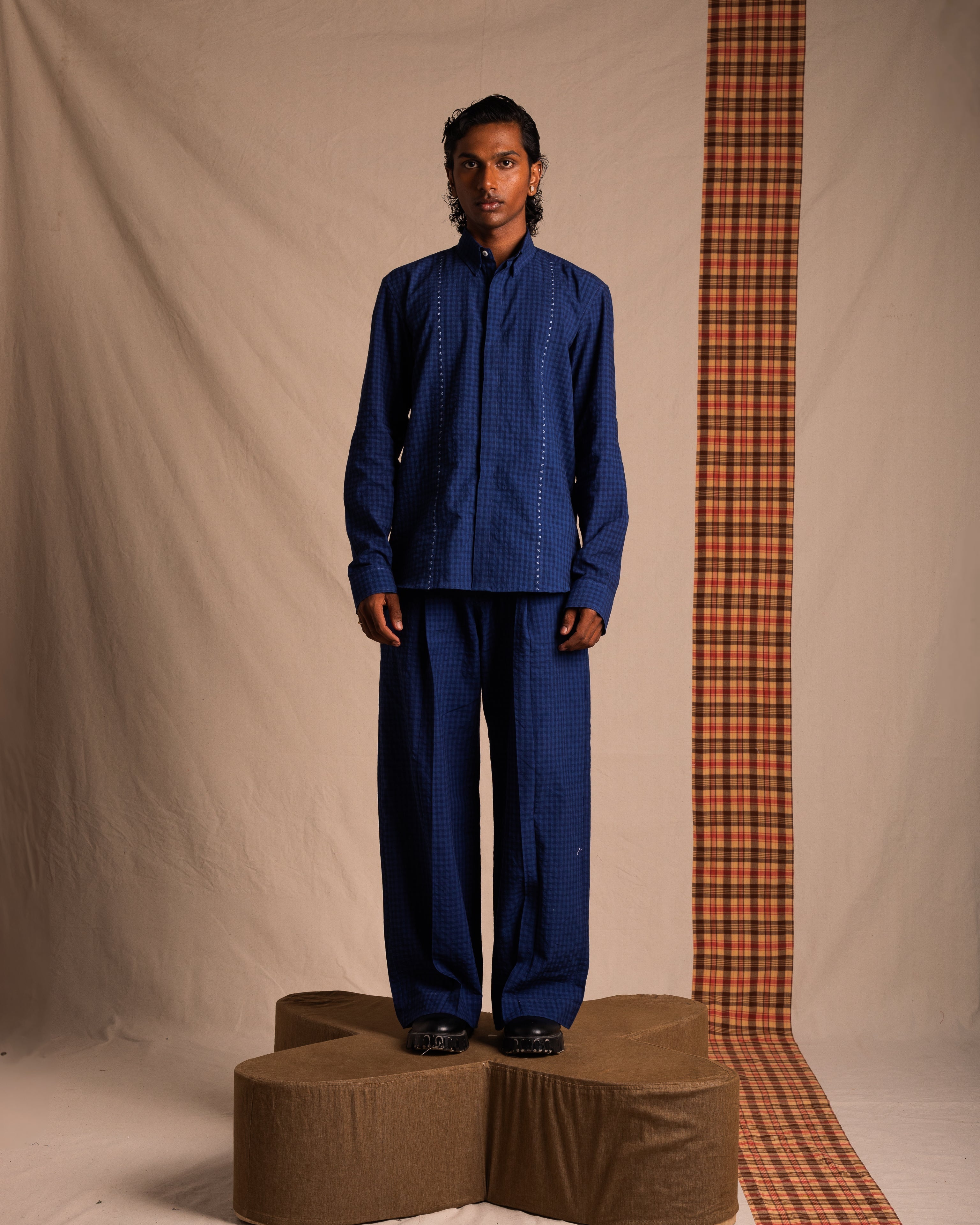 Man wearing blue checkered shirt and wide-leg pants, standing on brown platform against beige backdrop