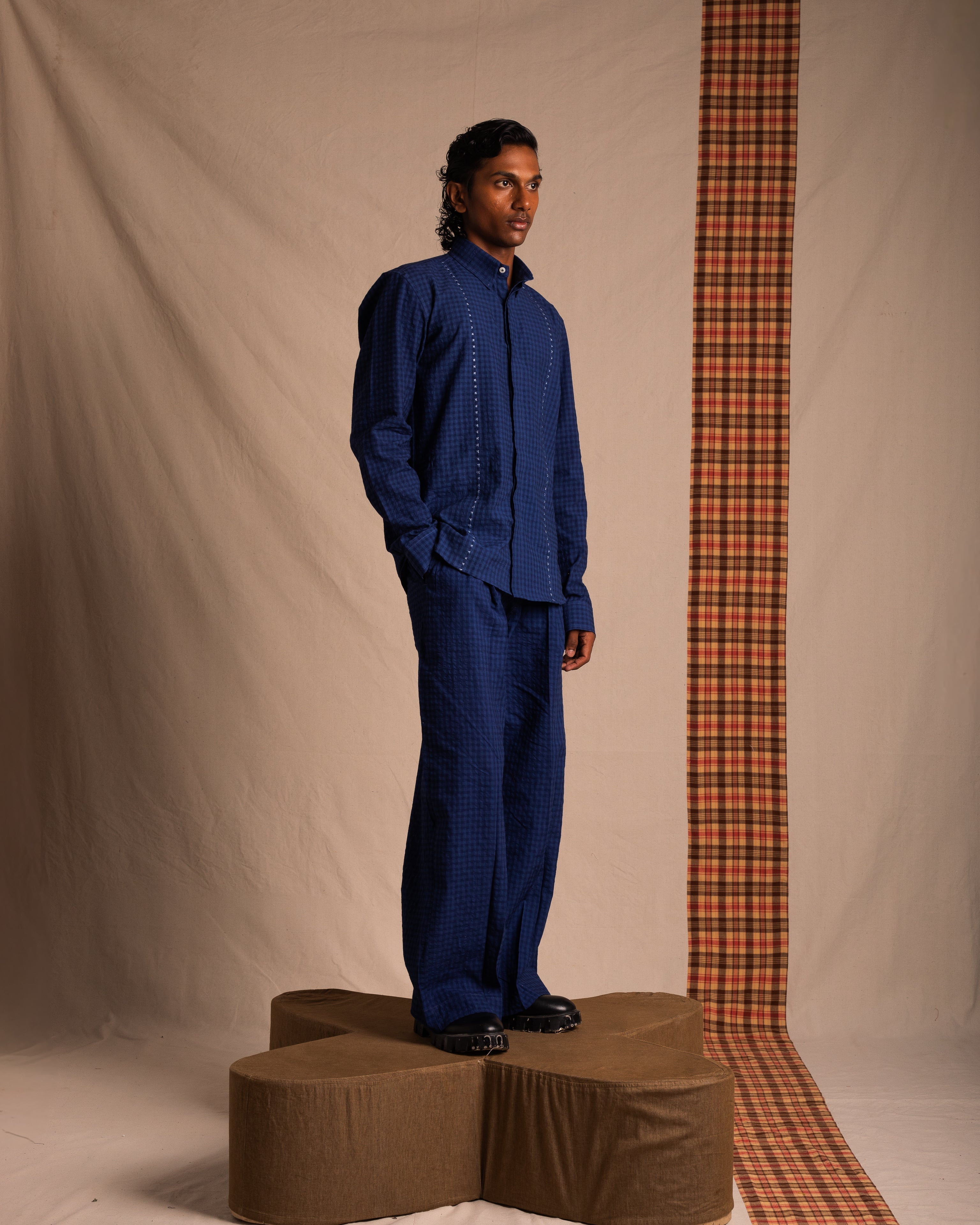 Man modeling blue checkered shirt and wide-leg pants, standing on geometric pedestal