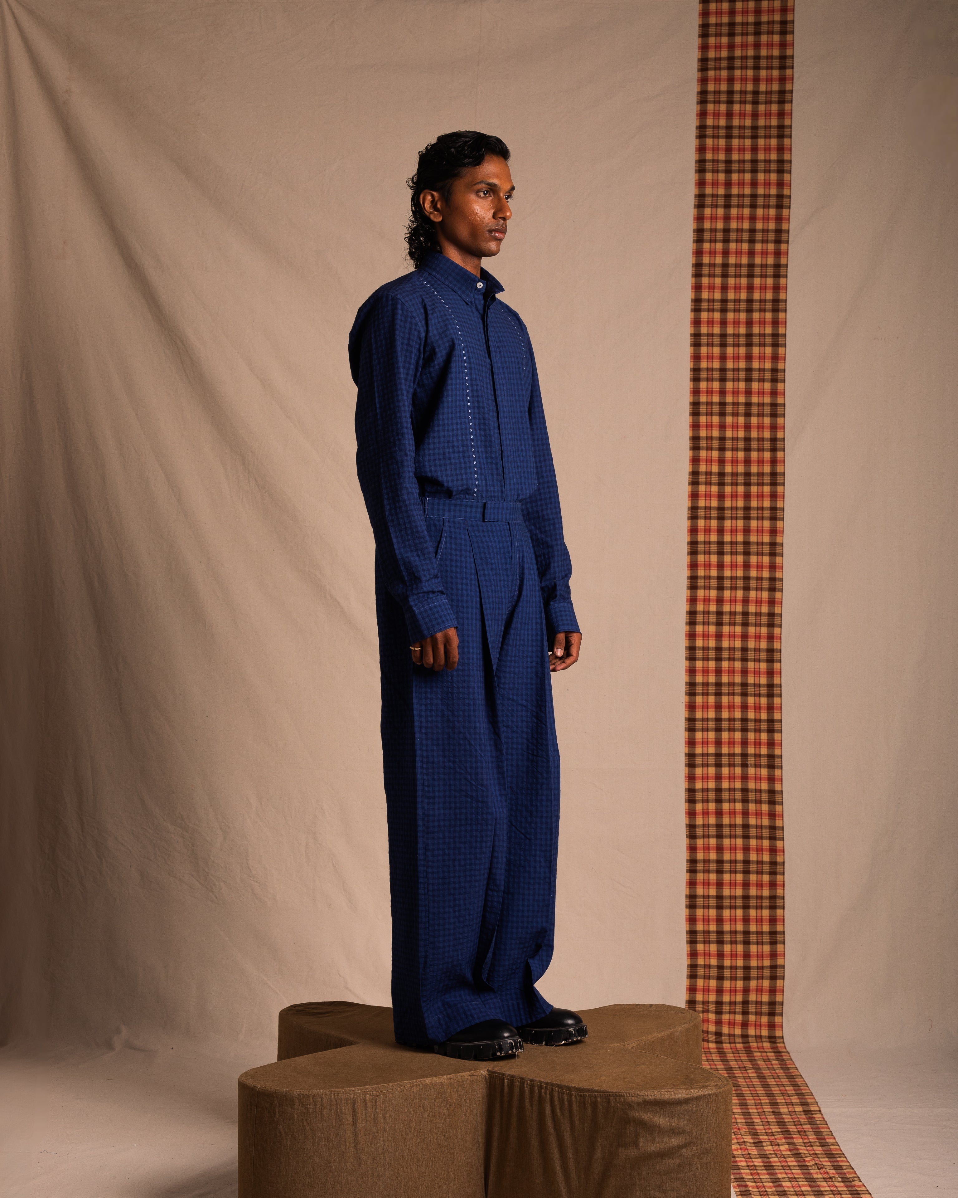 Man in loose blue checkered streetwear shirt and pants, standing in studio with plaid backdrop