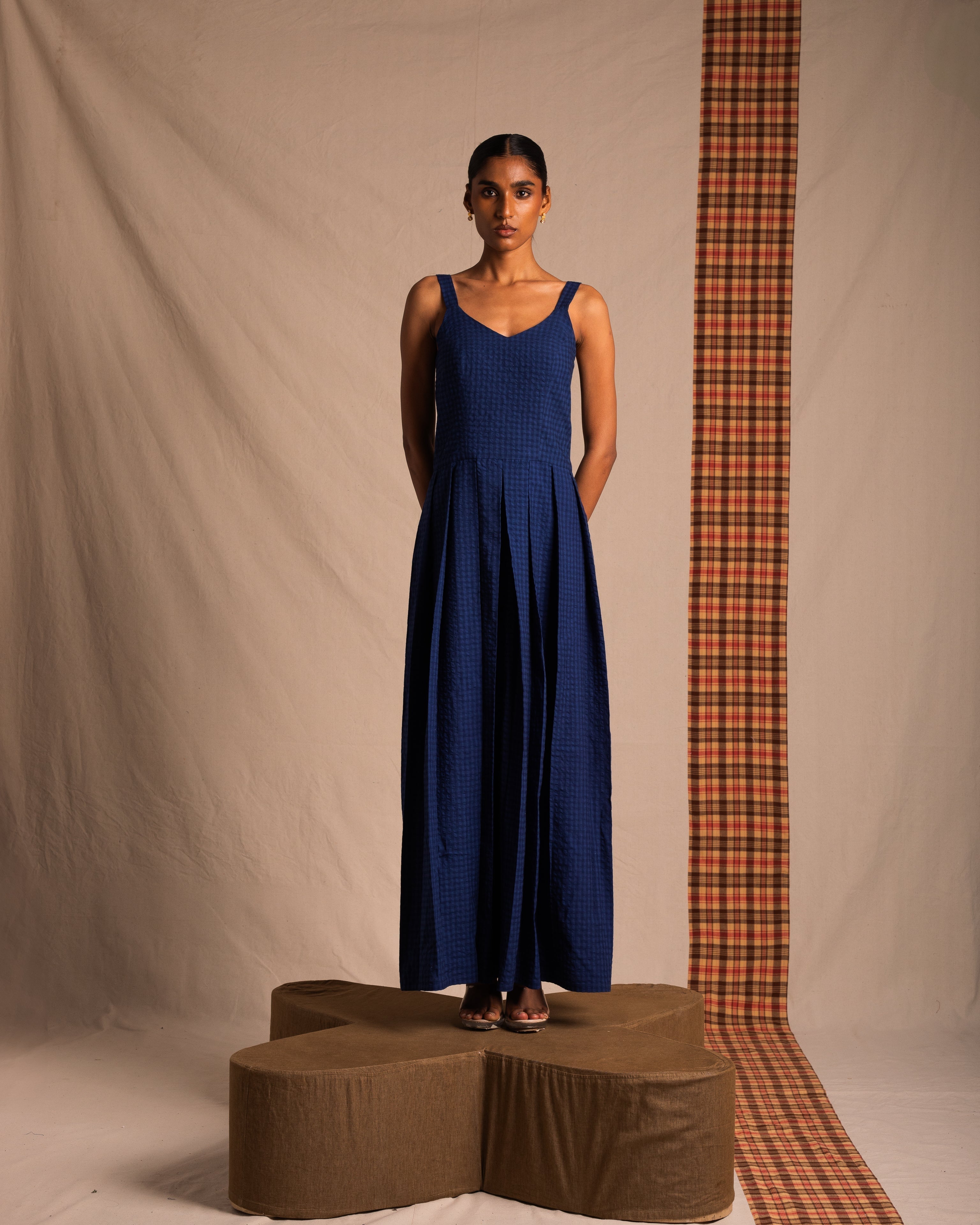 Woman modeling a sleeveless blue maxi dress, standing on a platform with a neutral and plaid backdrop.