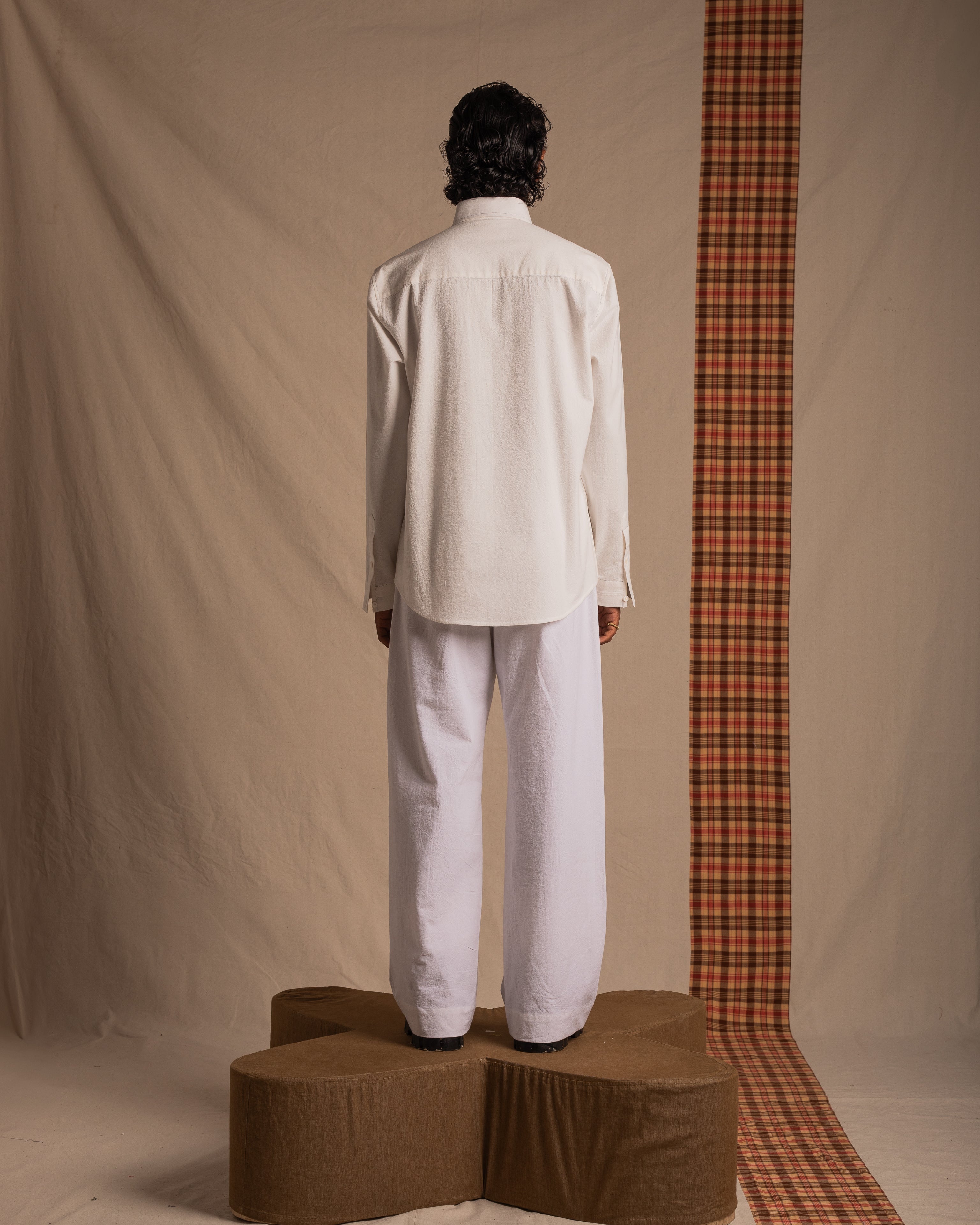 Man in white shirt and white pants stands on brown podium, back facing, neutral studio background